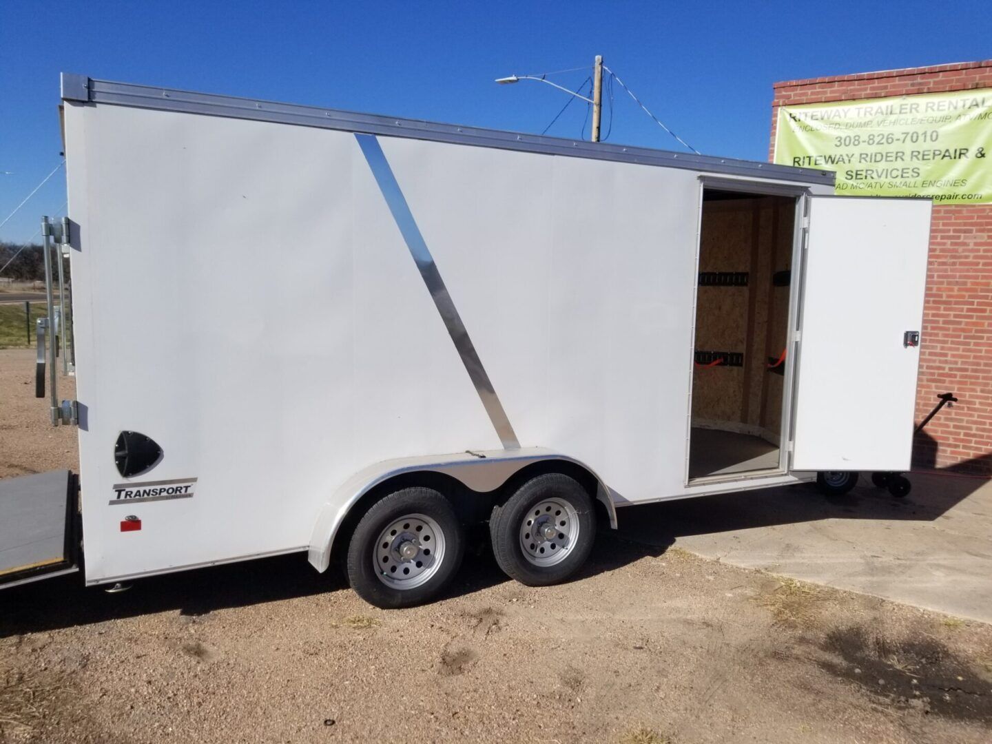White dual-axle enclosed trailer with open side door parked on a dirt lot under a clear blue sky.