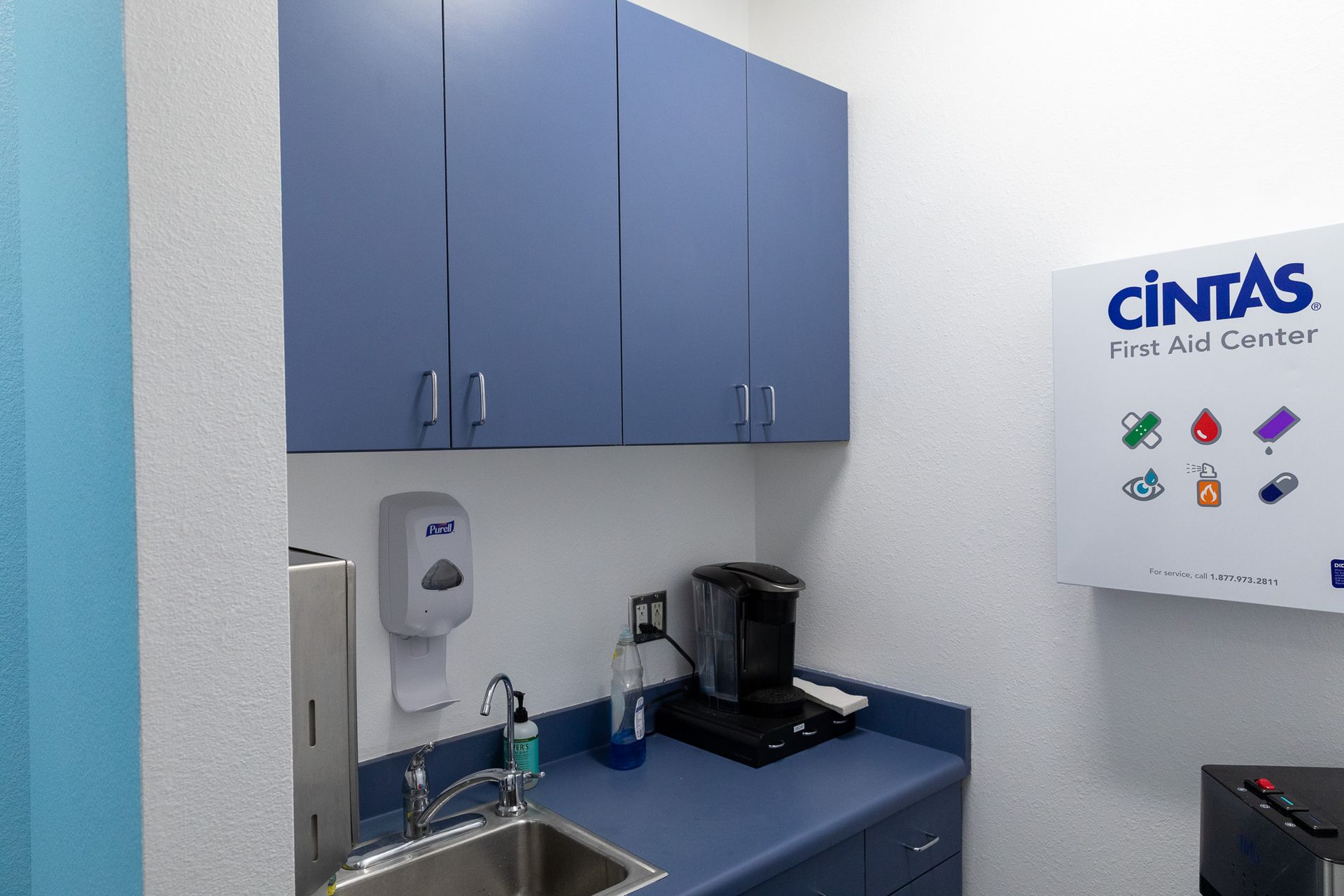 A small kitchenette with blue cabinets, a sink, and a coffee maker. A Cintas dispenser is mounted on the wall.
