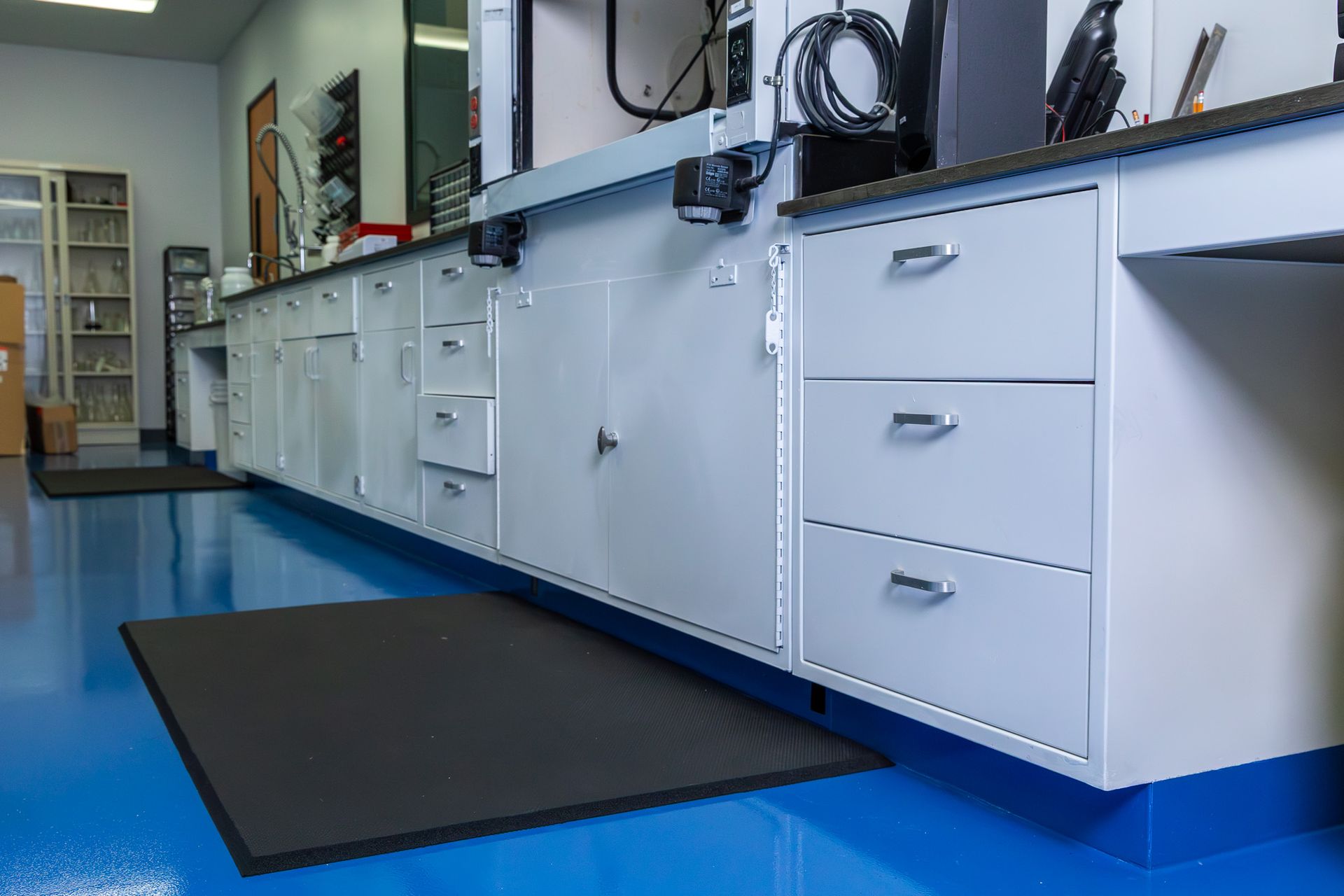 Blue-floored laboratory with white cabinets, drawers, and black mats.