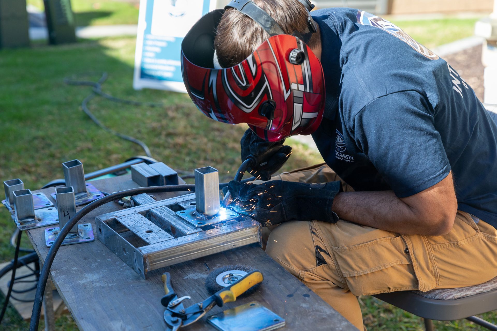 Welder in a red mask welds metal on a table outdoors; holding the torch with gloved hands.