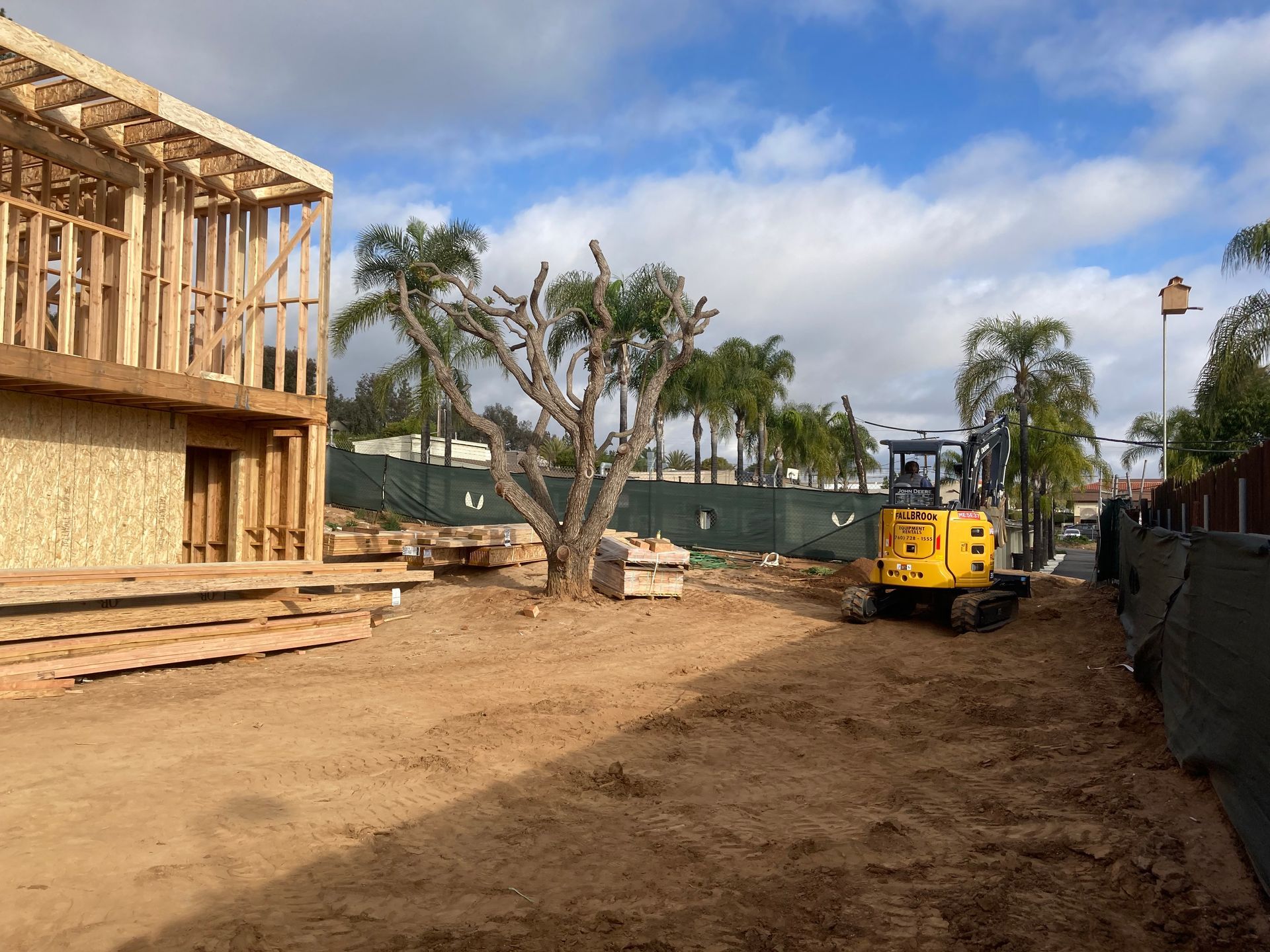 Construction site with a partially built wooden structure, excavator, and dirt ground under a cloudy sky.