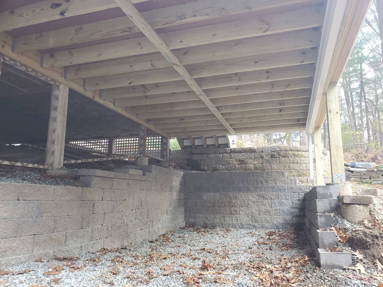 Underneath view of a deck supported by stone and concrete block foundation. Gravel and fallen leaves cover the ground.