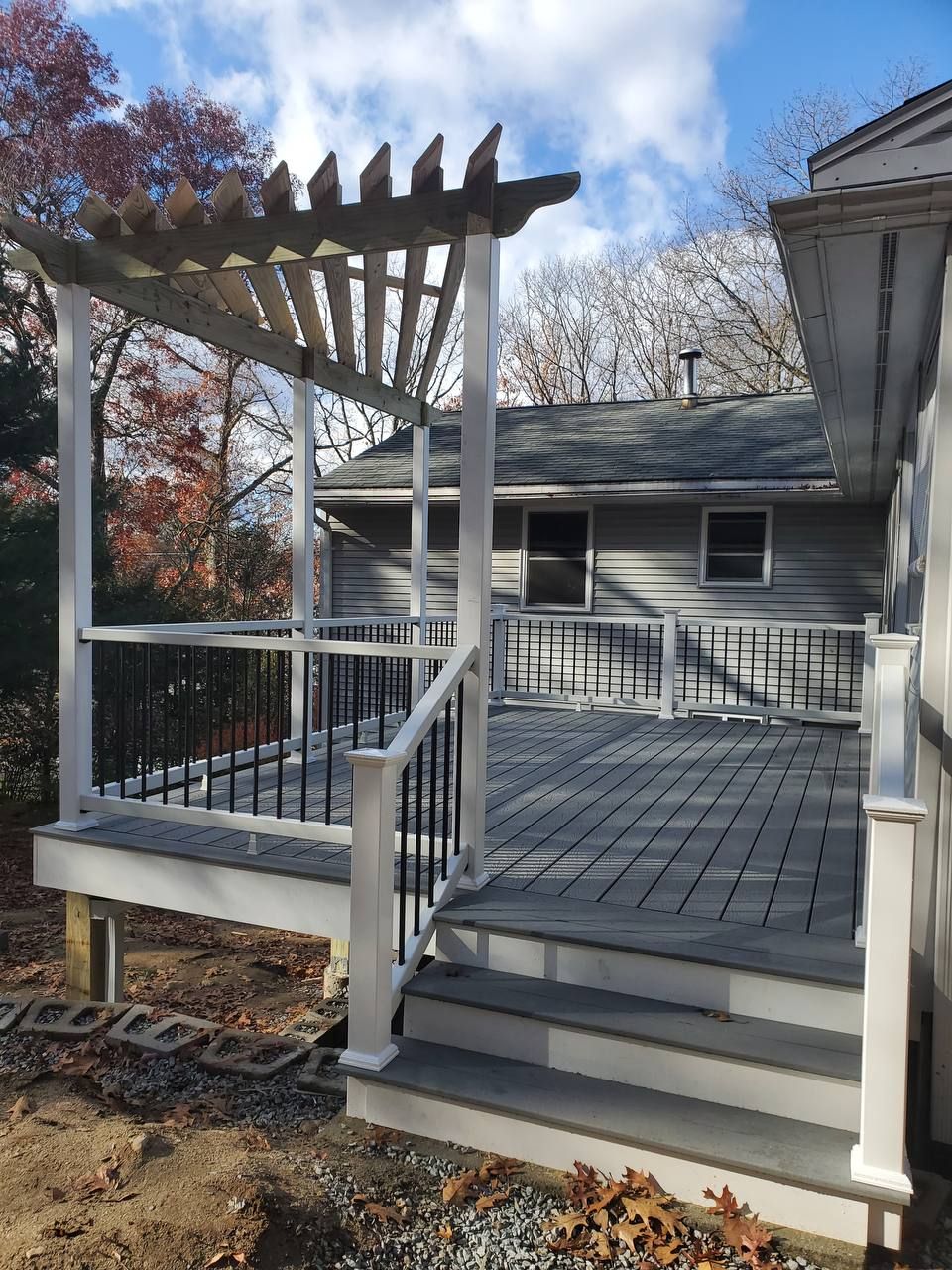 Wooden deck with pergola attached to a gray house, steps leading down, blue sky.
