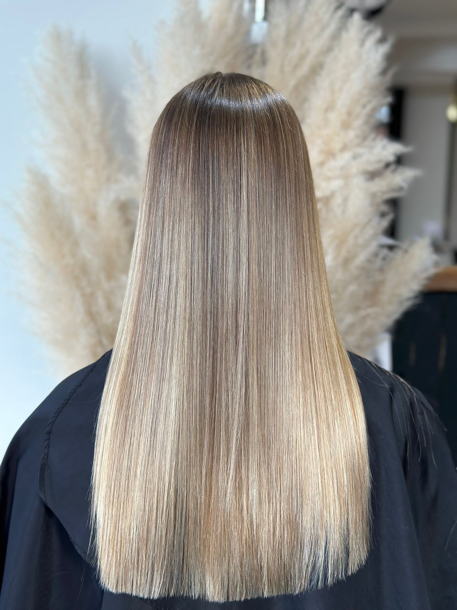 Long, straight, shiny hair transitioning from a dark brown root to a light blonde shade against a neutral background.
