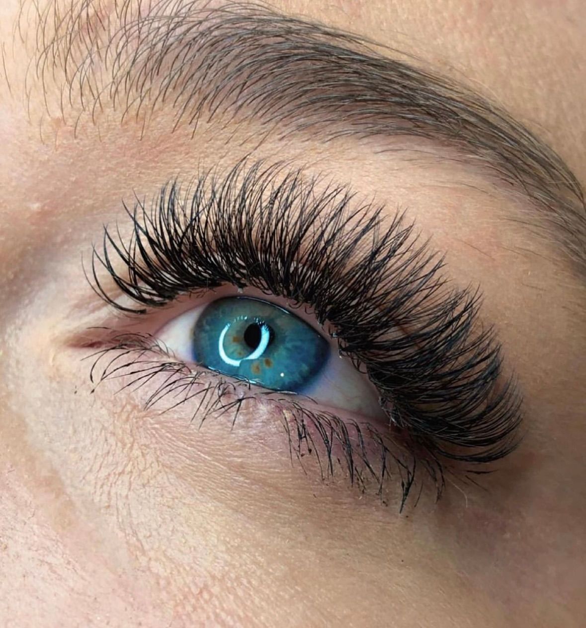 A close-up of a blue eye with long, dark, full eyelash extensions and a neatly groomed eyebrow.