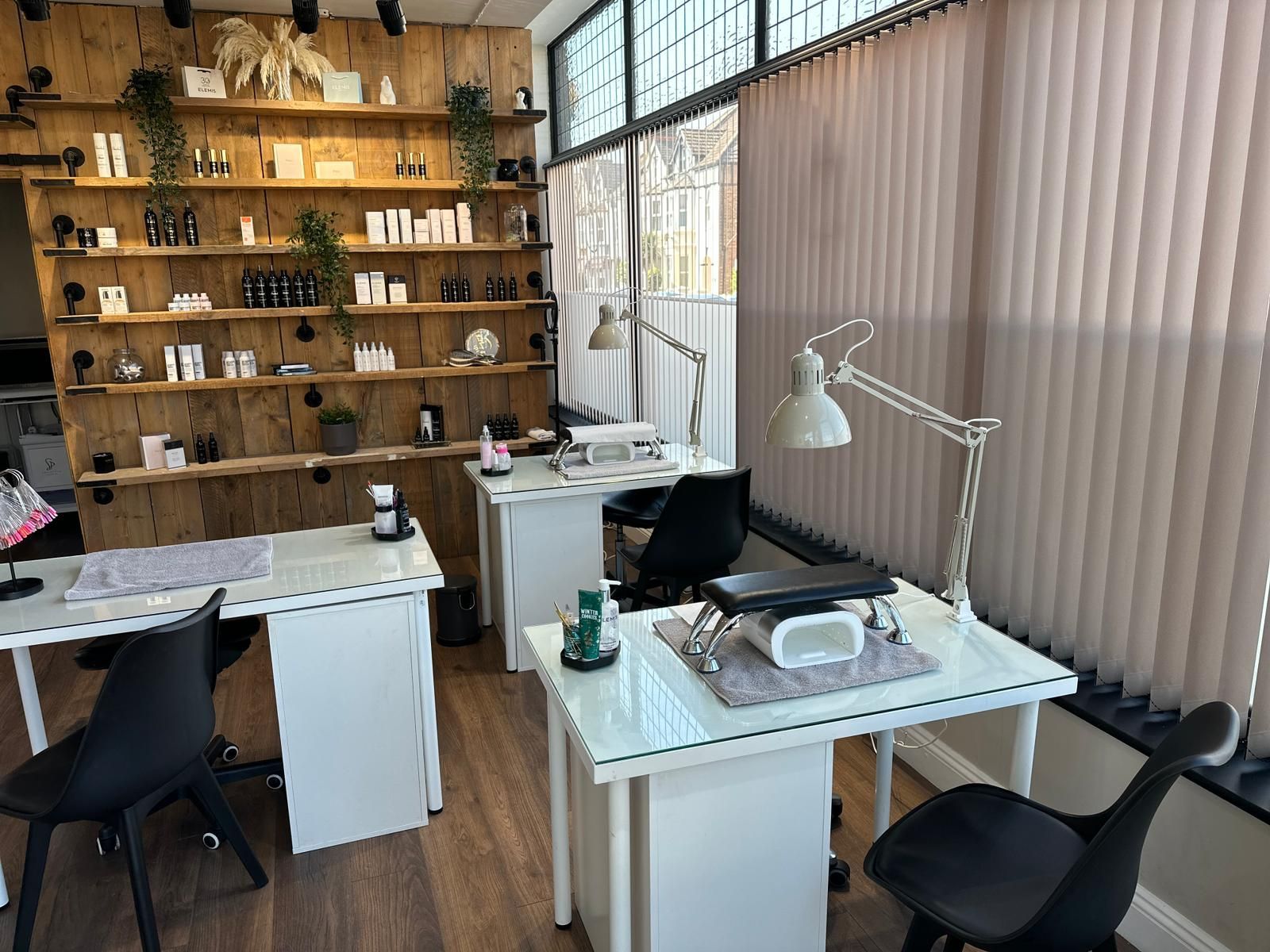A bright salon interior with two white manicure stations, task lamps, black chairs, and shelves stocked with products.
