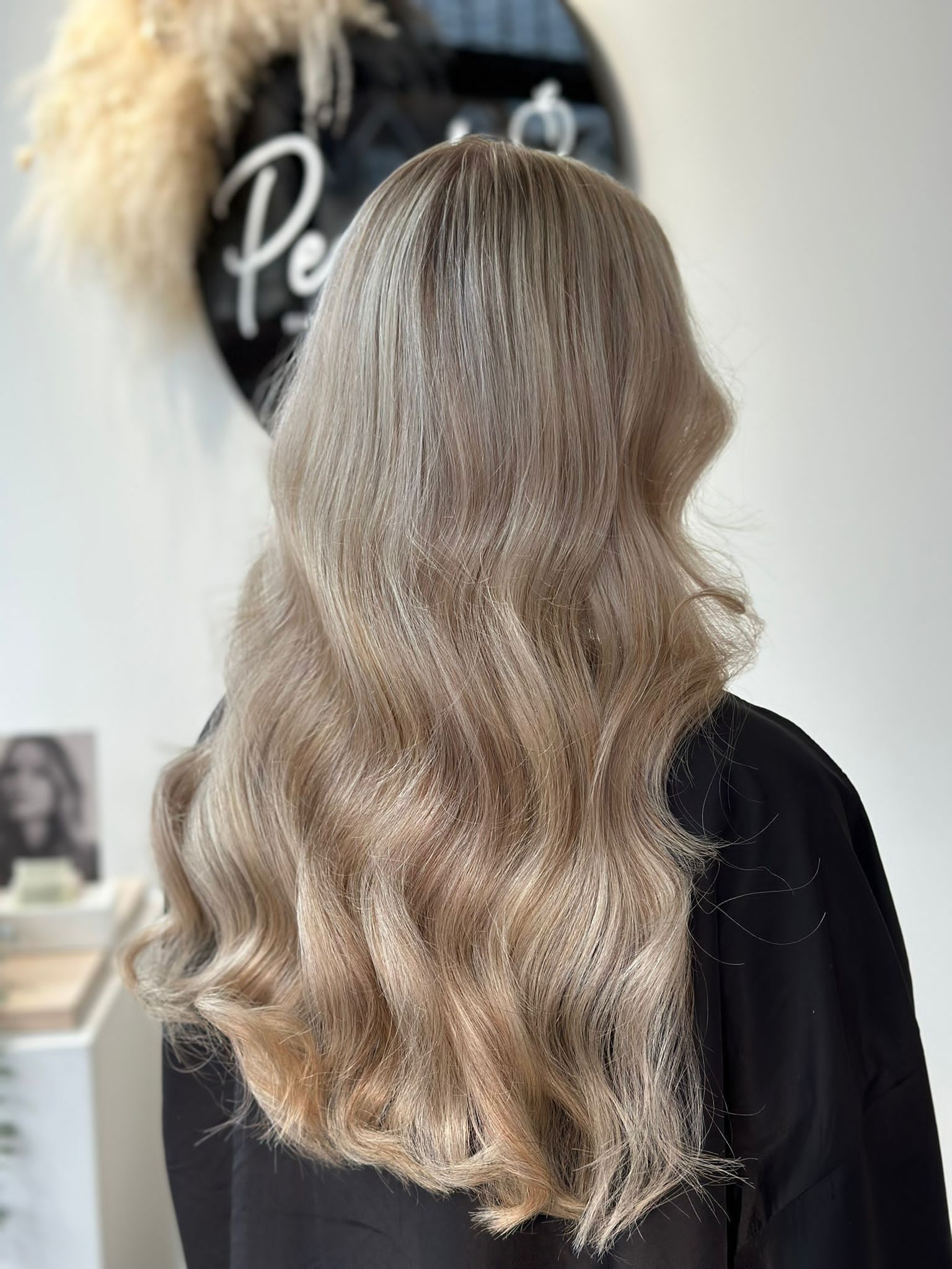 A person with long, light-brown wavy hair seen from behind in a salon setting.