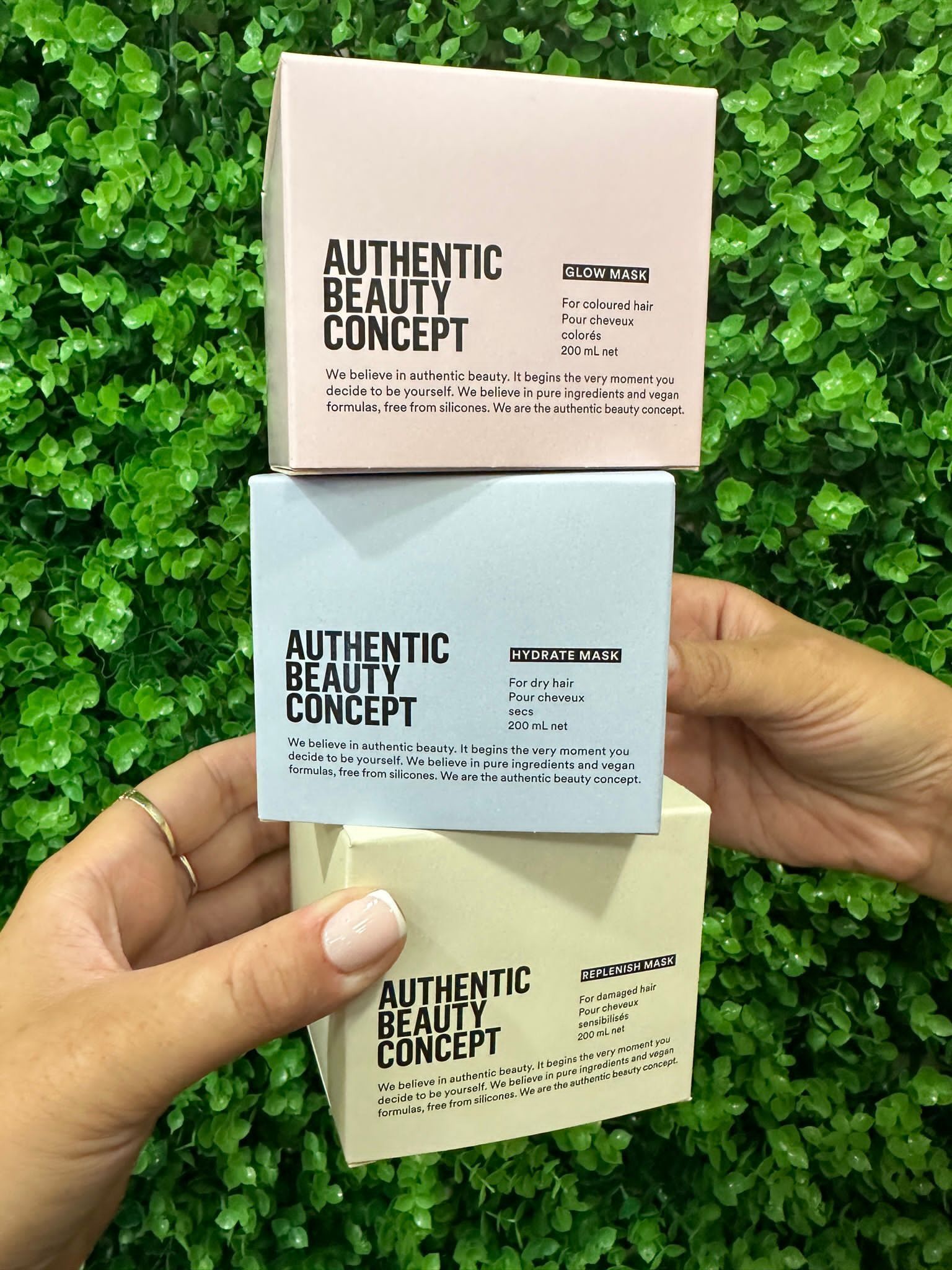 Two hands hold three stacked Authentic Beauty Concept product boxes in pink, blue, and cream against a green wall.