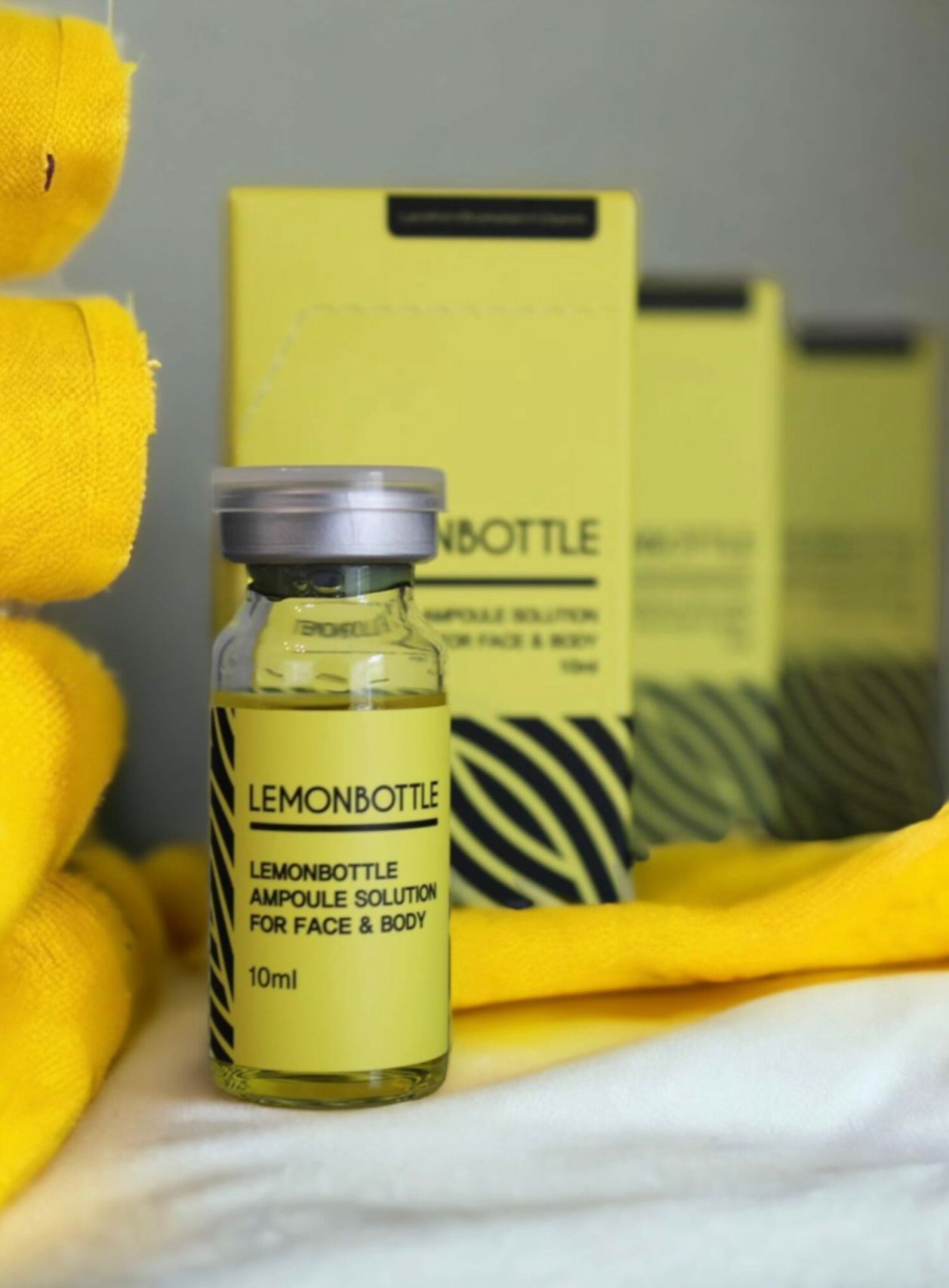 A glass vial and boxes of LEMONBOTTLE ampoule solution in bright yellow packaging, set against soft yellow fabric.