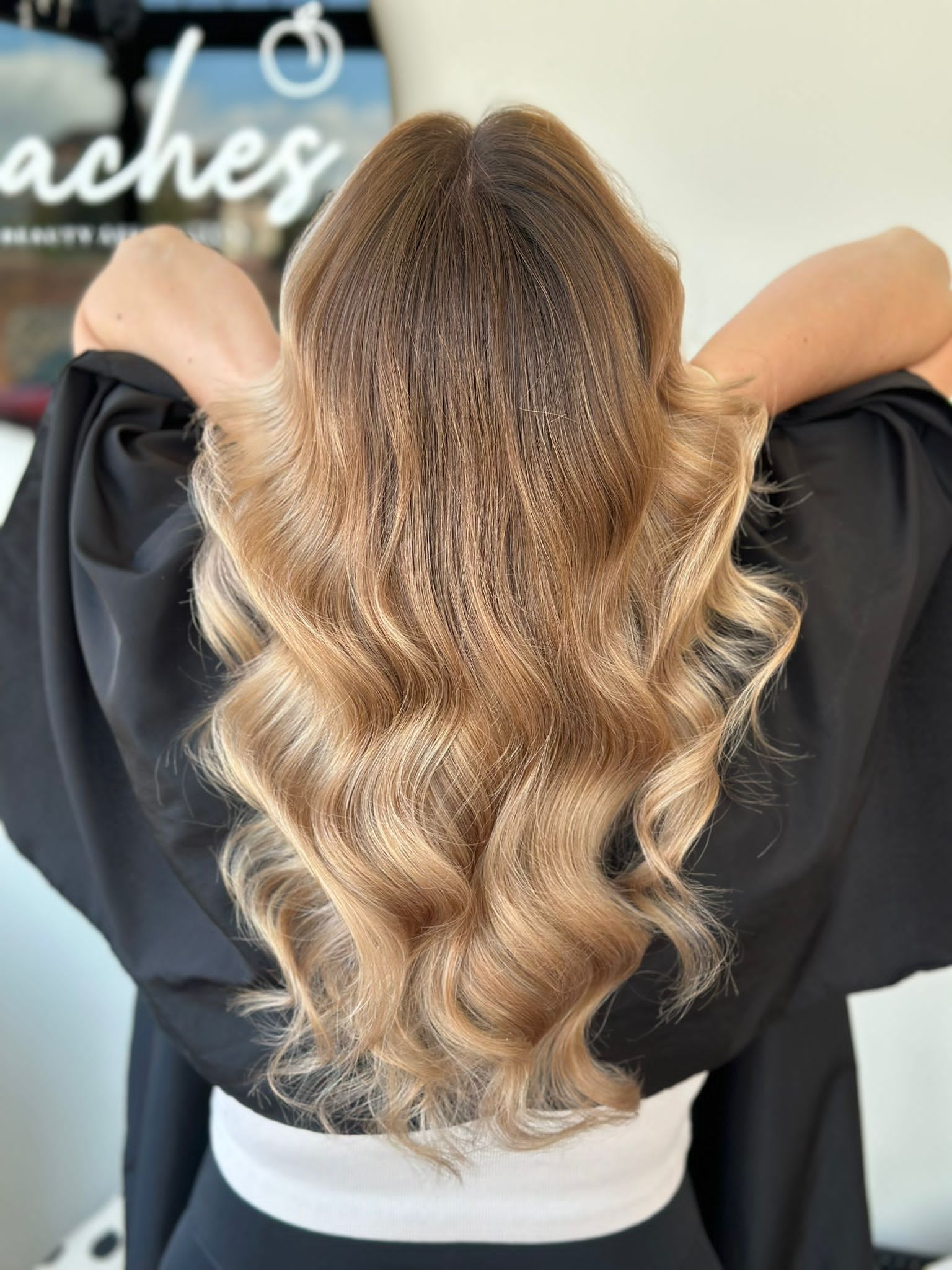 A person from behind shows long, wavy, golden-brown hair with a subtle crimped texture, styled in a salon.