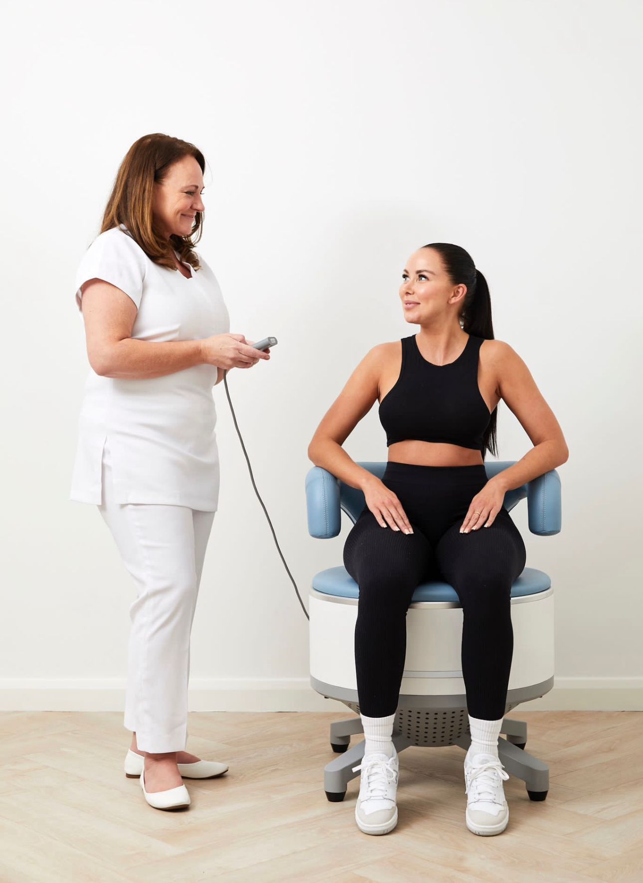 A practitioner holds a controller while a patient sits on a clinical therapy chair in a bright, minimalist room.