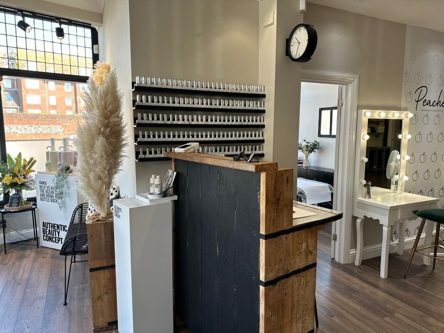 A bright beauty salon interior with a reception desk, nail polish shelves, a makeup station with vanity lights, and plants.
