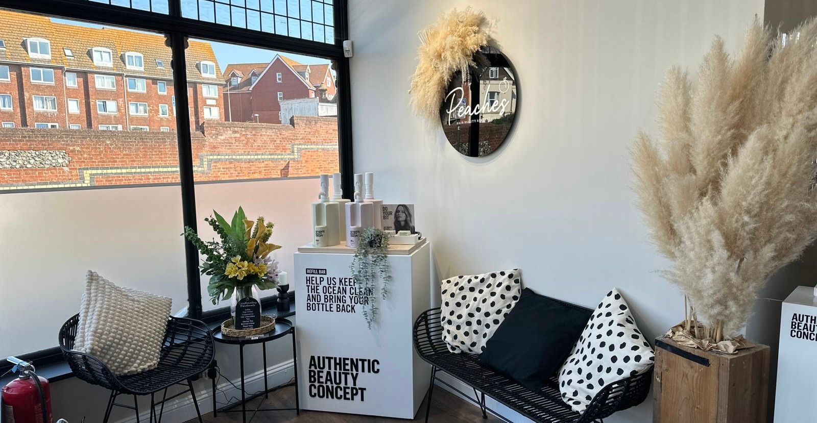 A modern salon waiting area with a black wire chair, a bench with patterned pillows, product displays, and dried plants.