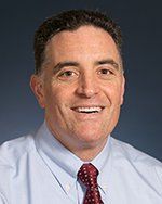 A man in a blue shirt and red tie is smiling for the camera.