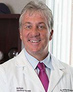 A man in a white coat and pink tie is smiling for the camera.