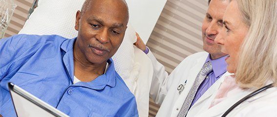A man in a hospital bed is looking at a tablet with two doctors.