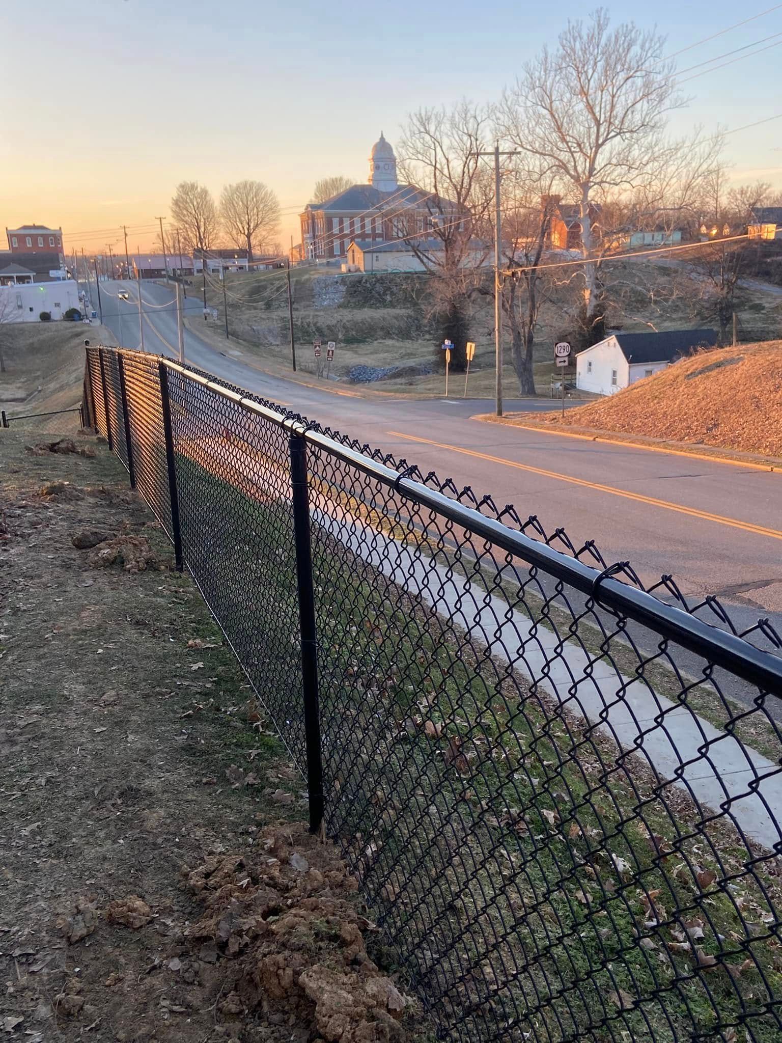 Black chain-link fence lines a grassy slope next to a road, with a building and trees in the background.