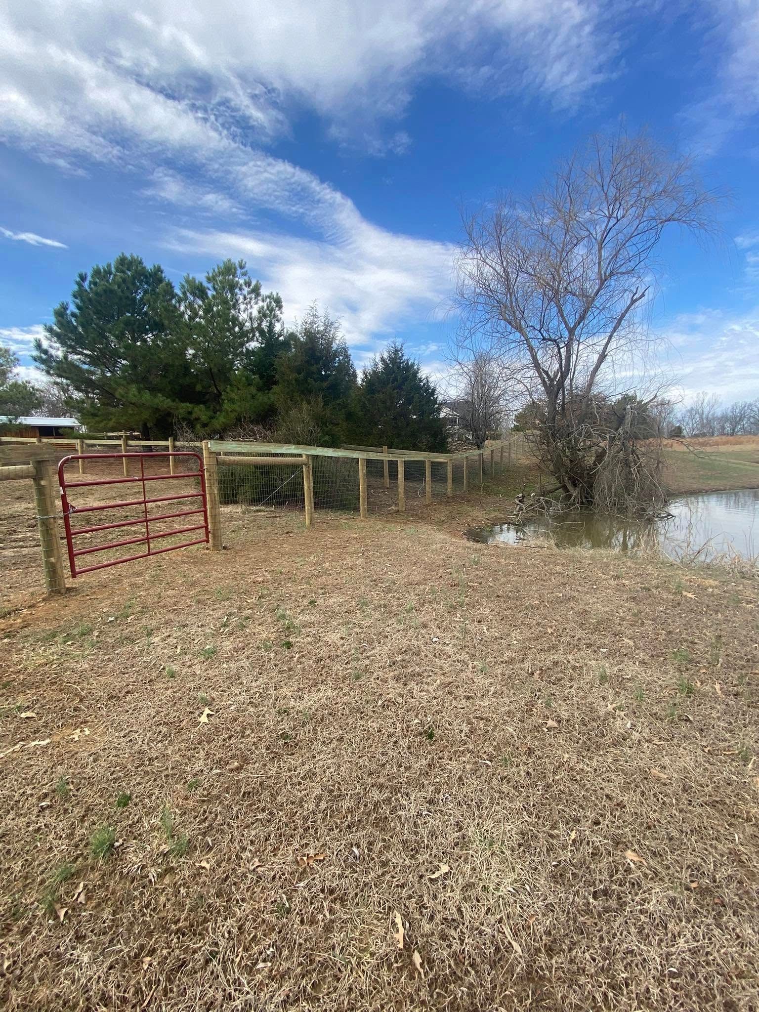 A wooden fence surrounds a brown, dry field, with a small body of water on the right and a blue sky above.