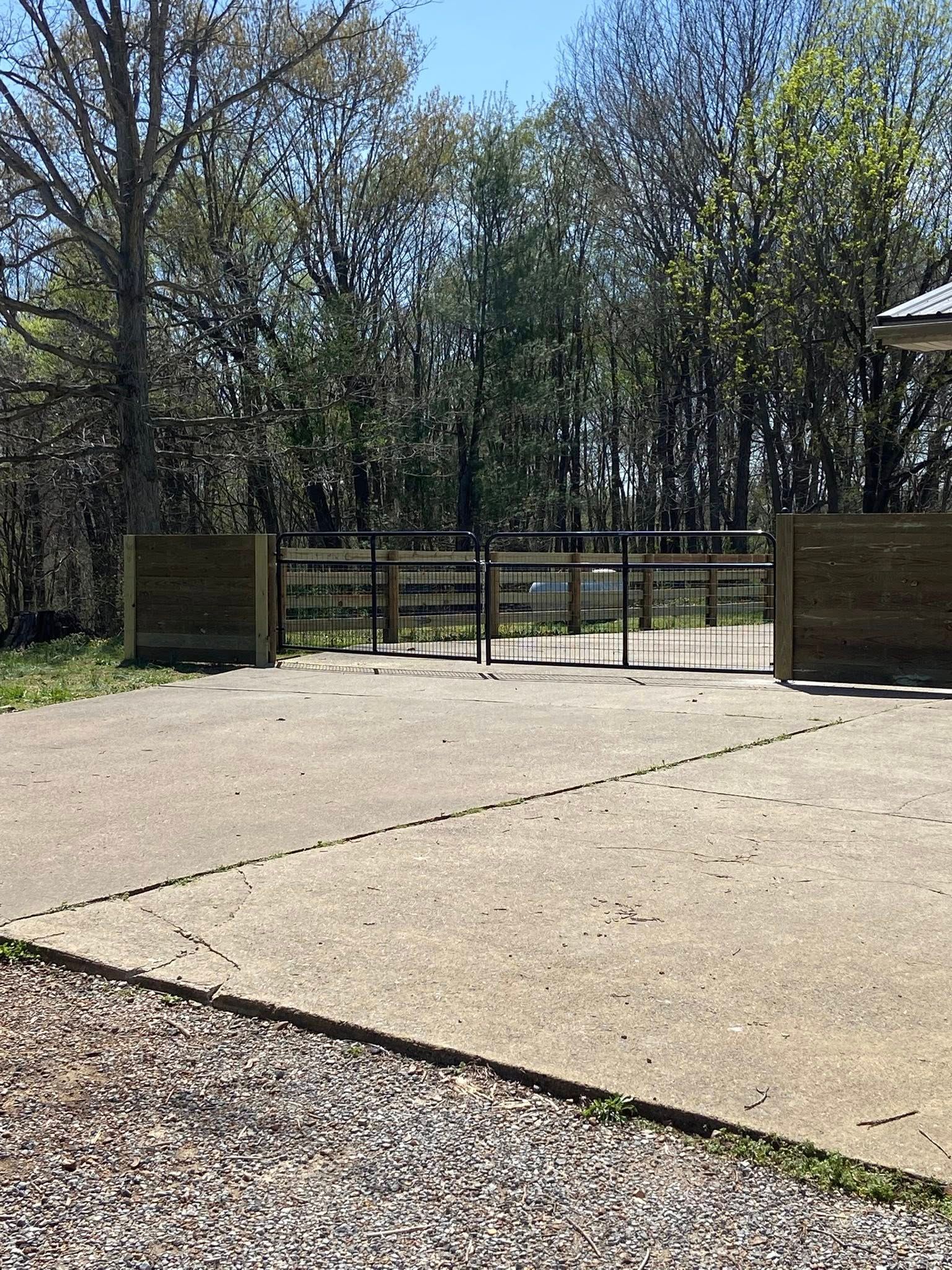 Concrete drive leads to black metal gate, flanked by wooden posts. Trees in background.