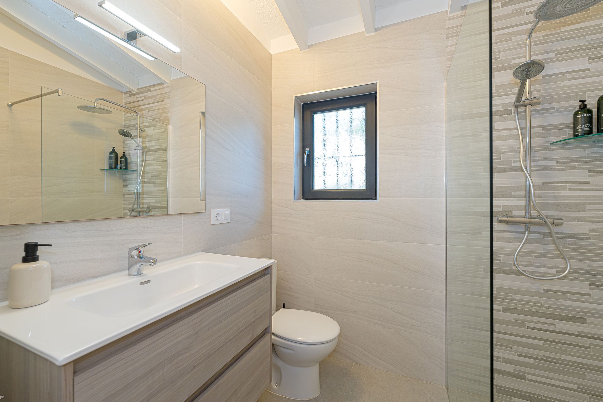 Moderne badkamer met wit badmeubel, grote spiegel, toilet, betegelde douche en klein raam.