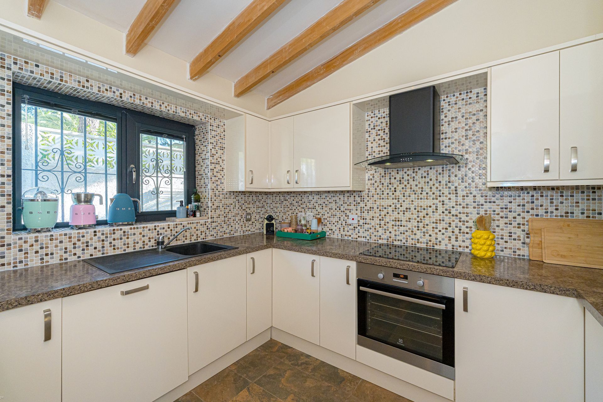 Moderne keuken met witte kasten, mozaïektegels als achterwand, zwarte apparaten en houten plafondbalken.