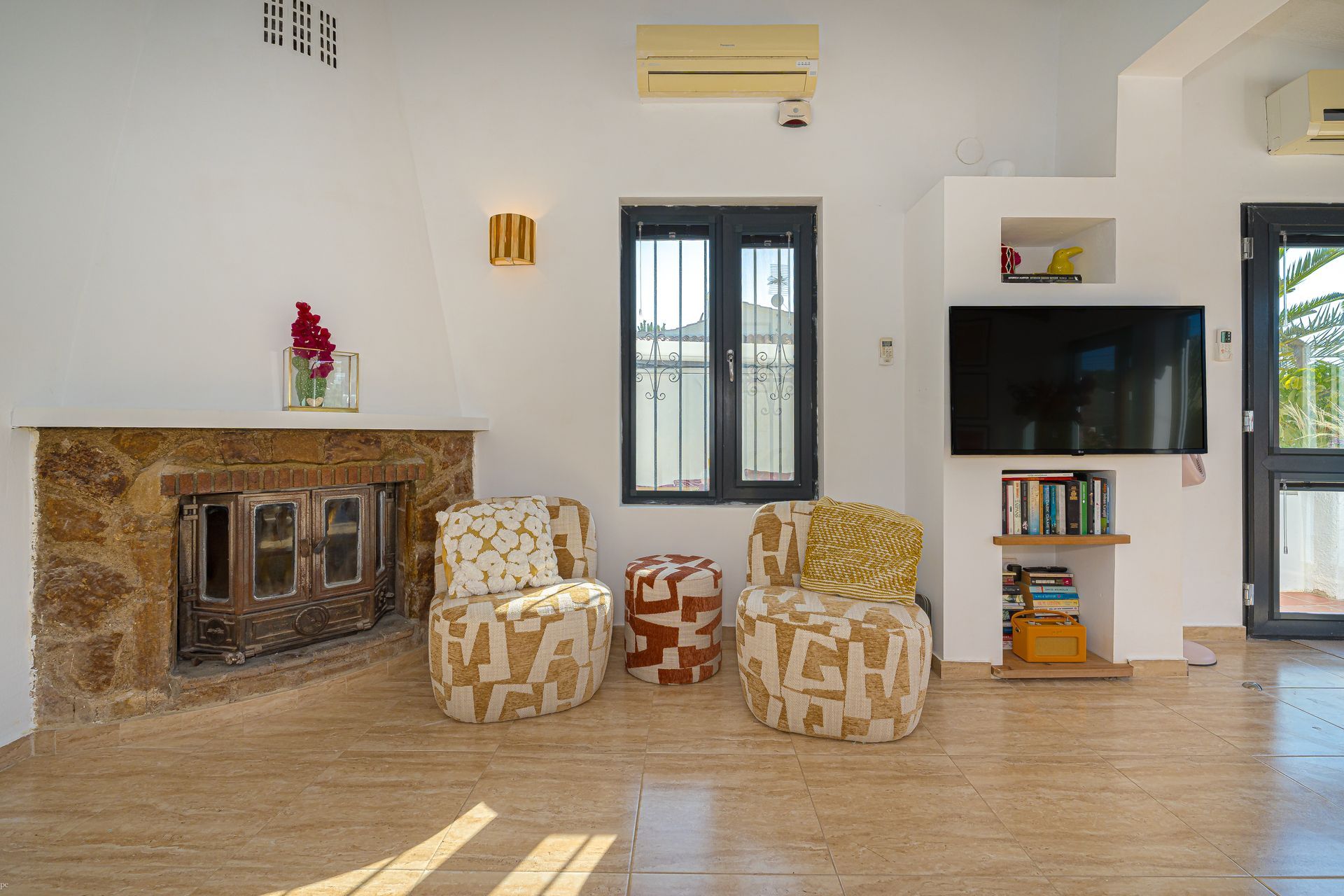 Lichte woonkamer met stenen open haard, stoelen met patroon, tv en openslaande deuren naar een patio.