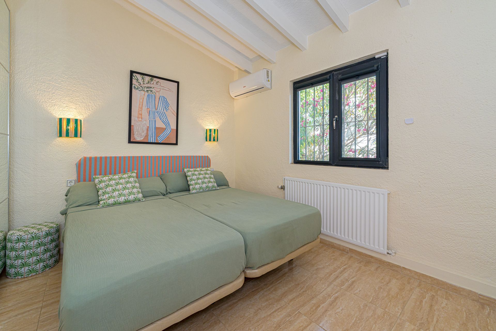 Minimalistische slaapkamer met twee eenpersoonsbedden, kussens met patronen, kleurrijke muurdecoratie en een raam met natuurlijk licht.