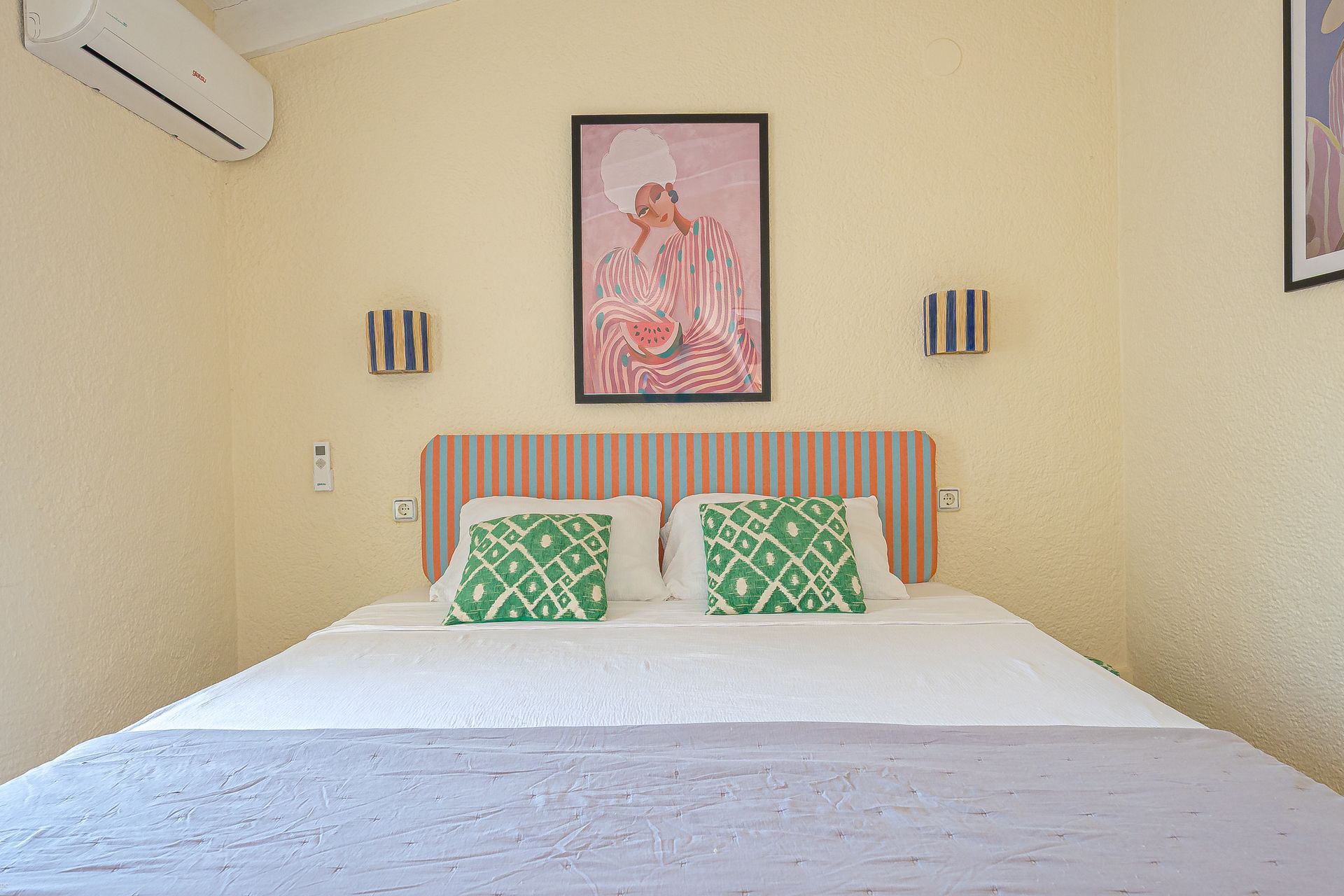 Lichte slaapkamer met een wit bed, groene kussens met patroon, een oranje gestreept hoofdbord en ingelijste kunst aan beige muren.