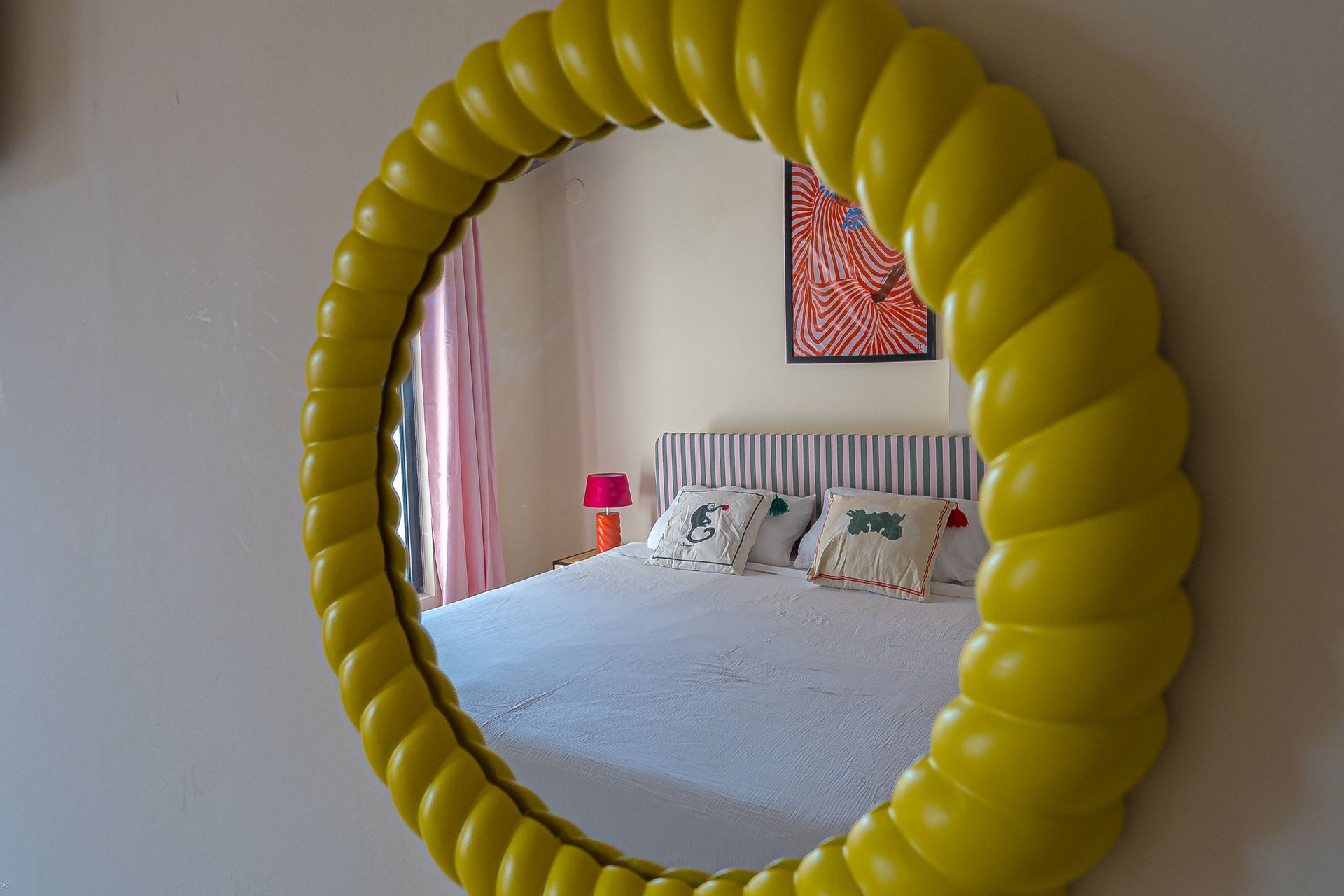 Een spiegel met een gele ballonvormige lijst weerspiegelt een lichte slaapkamer met een bed en roze gordijnen.