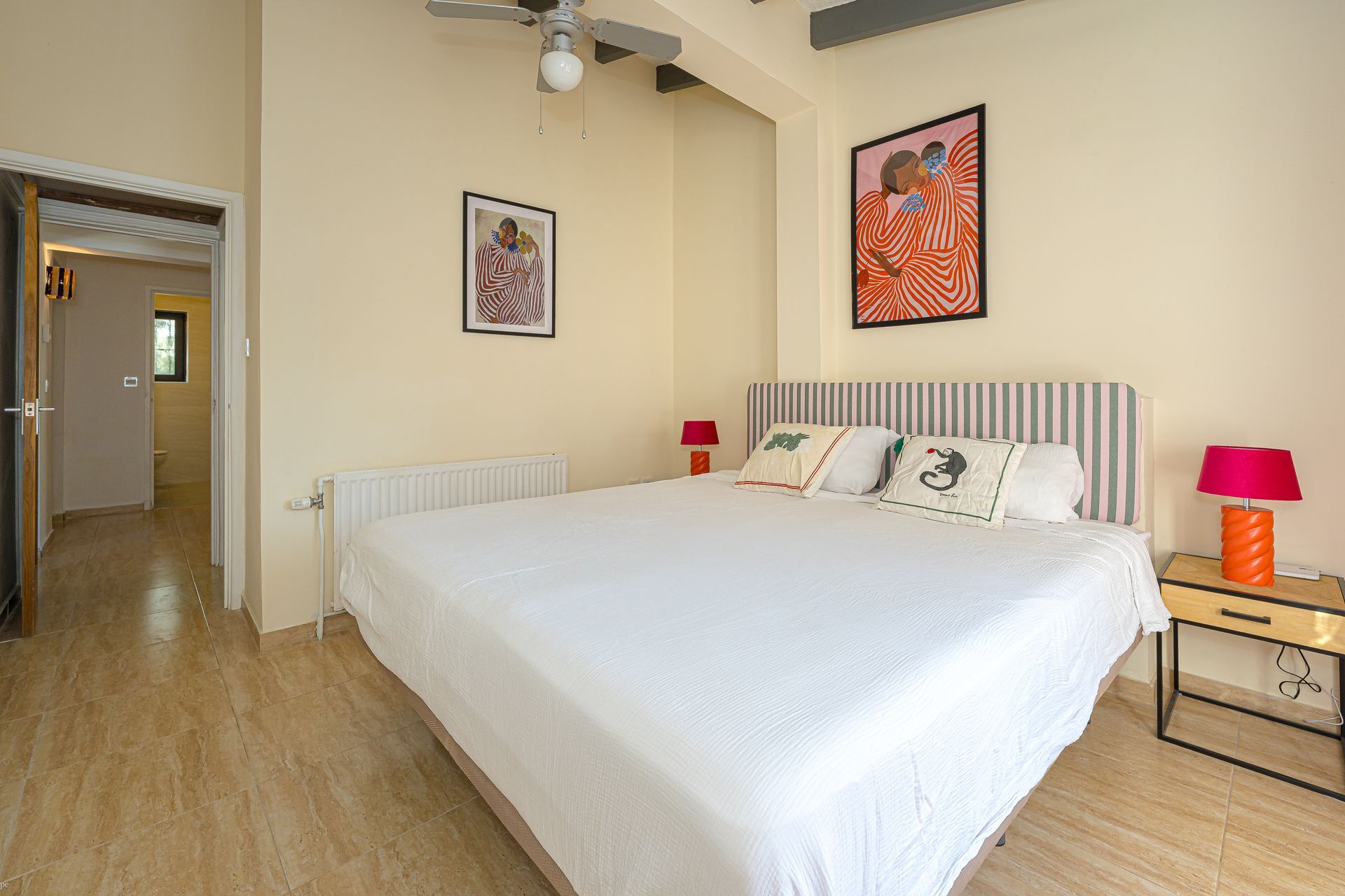 Lichte slaapkamer met een groot wit bed, een gestreept hoofdbord, roze lampen en ingelijste kunstwerken aan de muur.