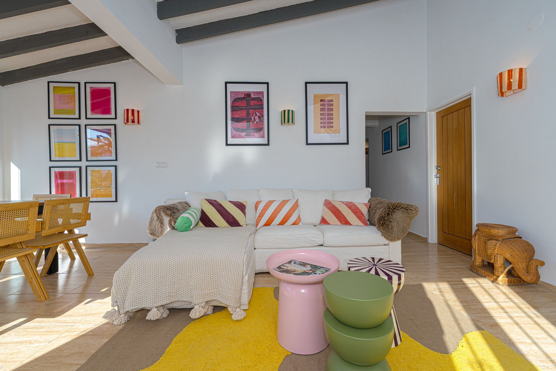 Lichte woonkamer met gestreepte bank, kleurrijke muurdecoratie, gevlochten stoelen en roze en groene krukjes in het zonlicht.
