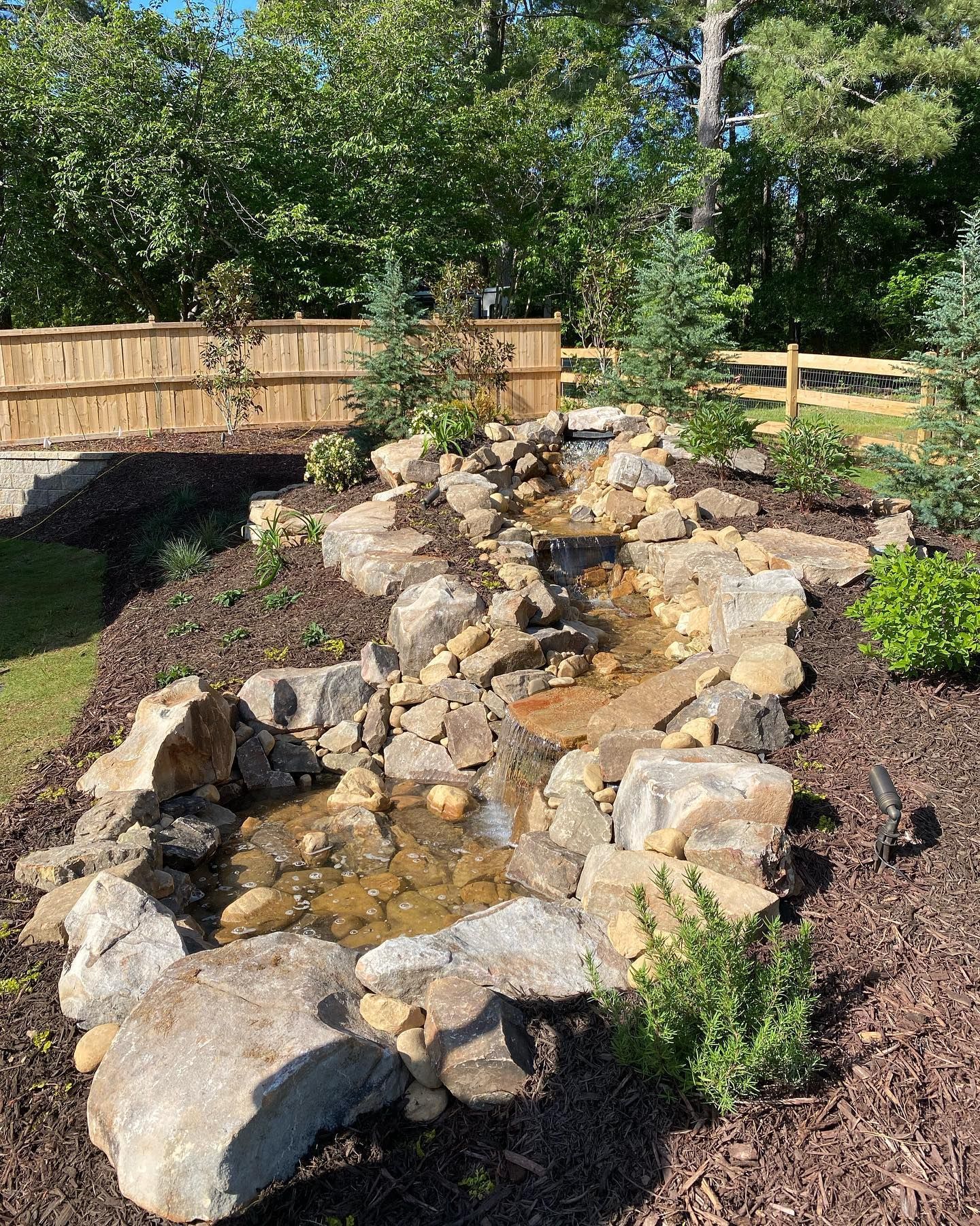 Rock garden waterfall with large boulders, mulch, and a wooden fence in a sunny yard