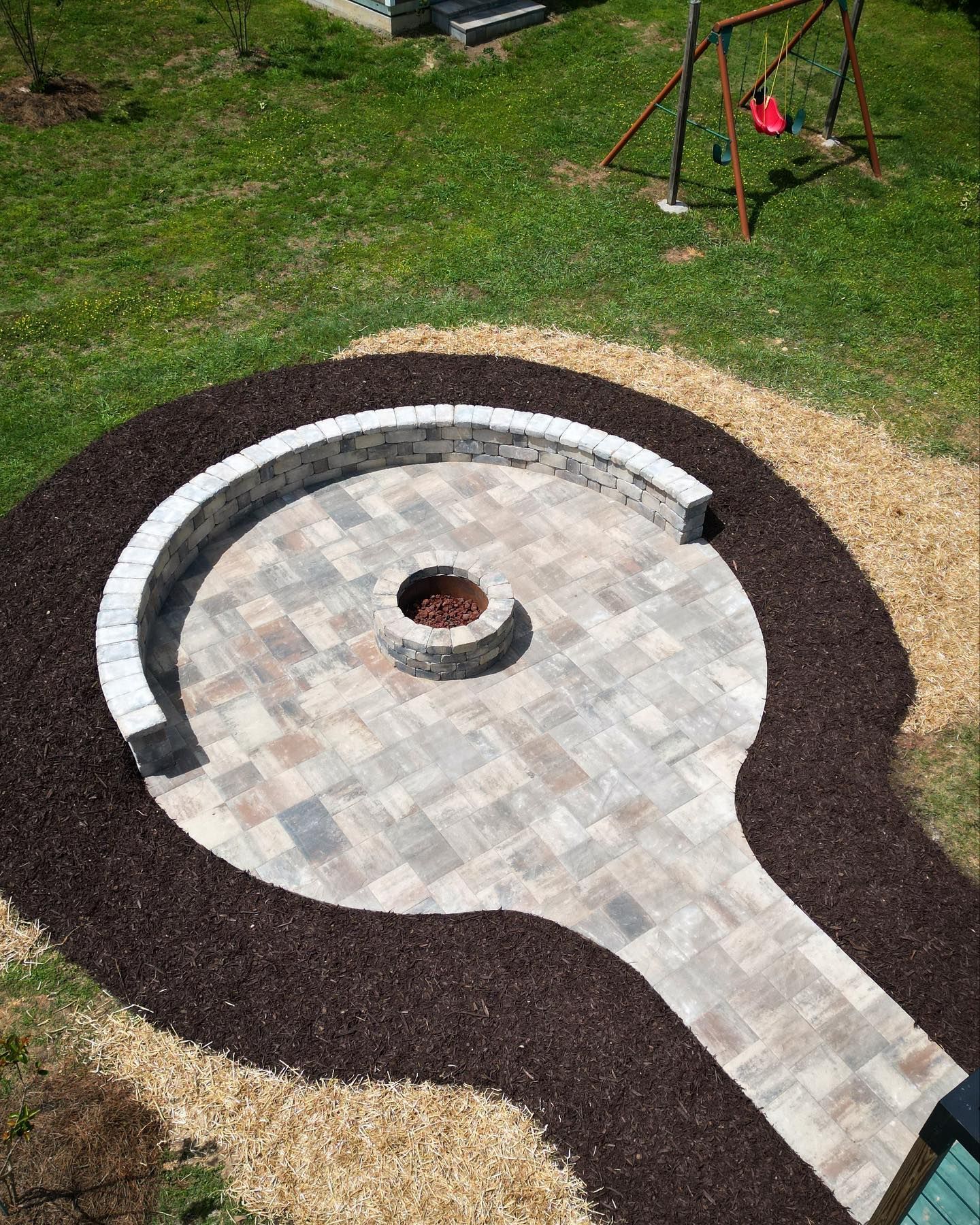 Curved stone patio with a central fire pit, bordered by dark mulch and gravel in a backyard lawn.