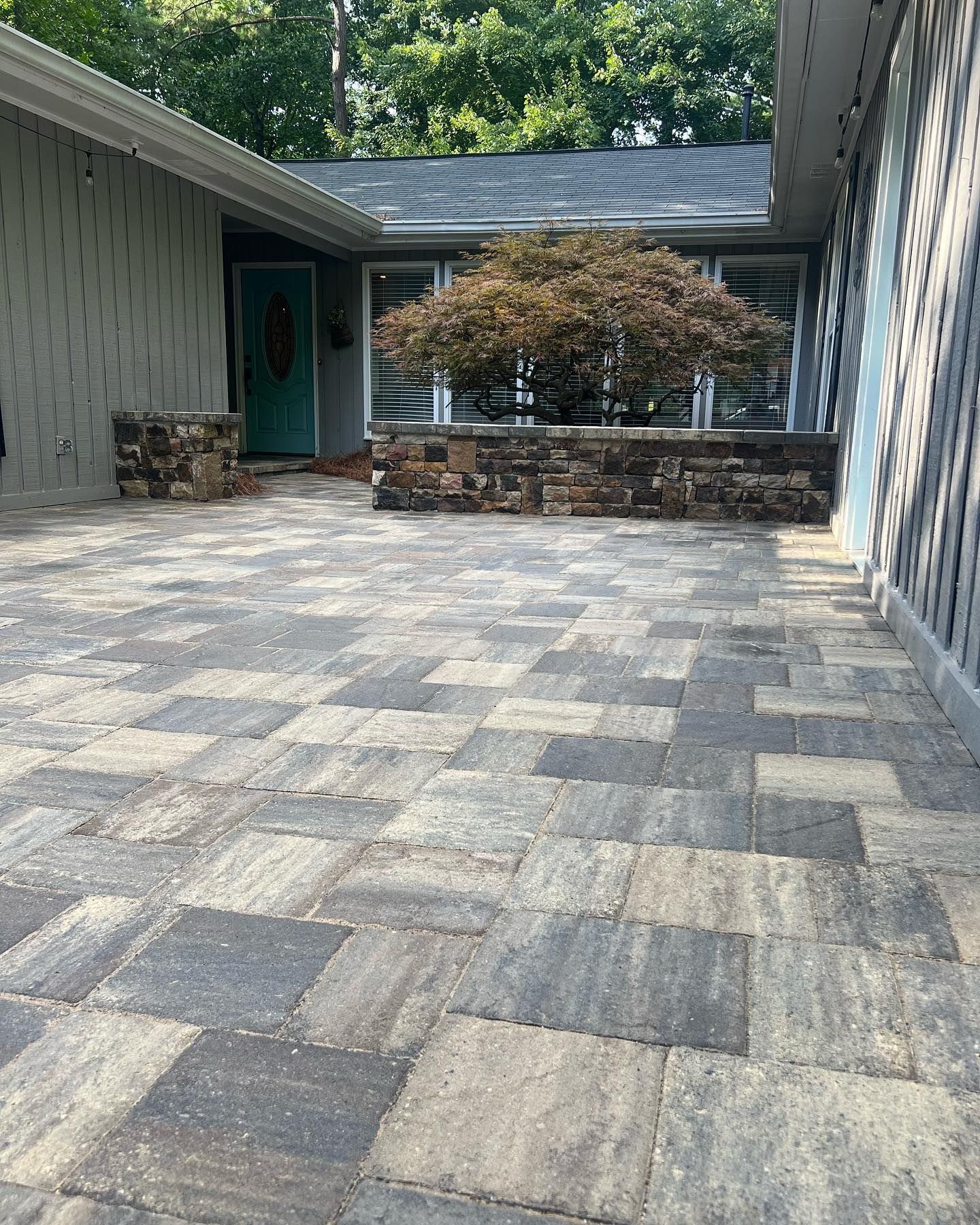 Stone patio walkway beside a house, with gray pavers, a teal door, and a stone planter under trees