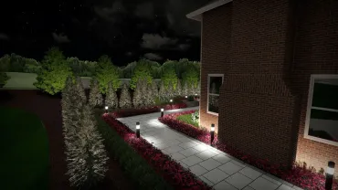Illuminated garden path beside a brick house at night, lined with hedges and red mulch.