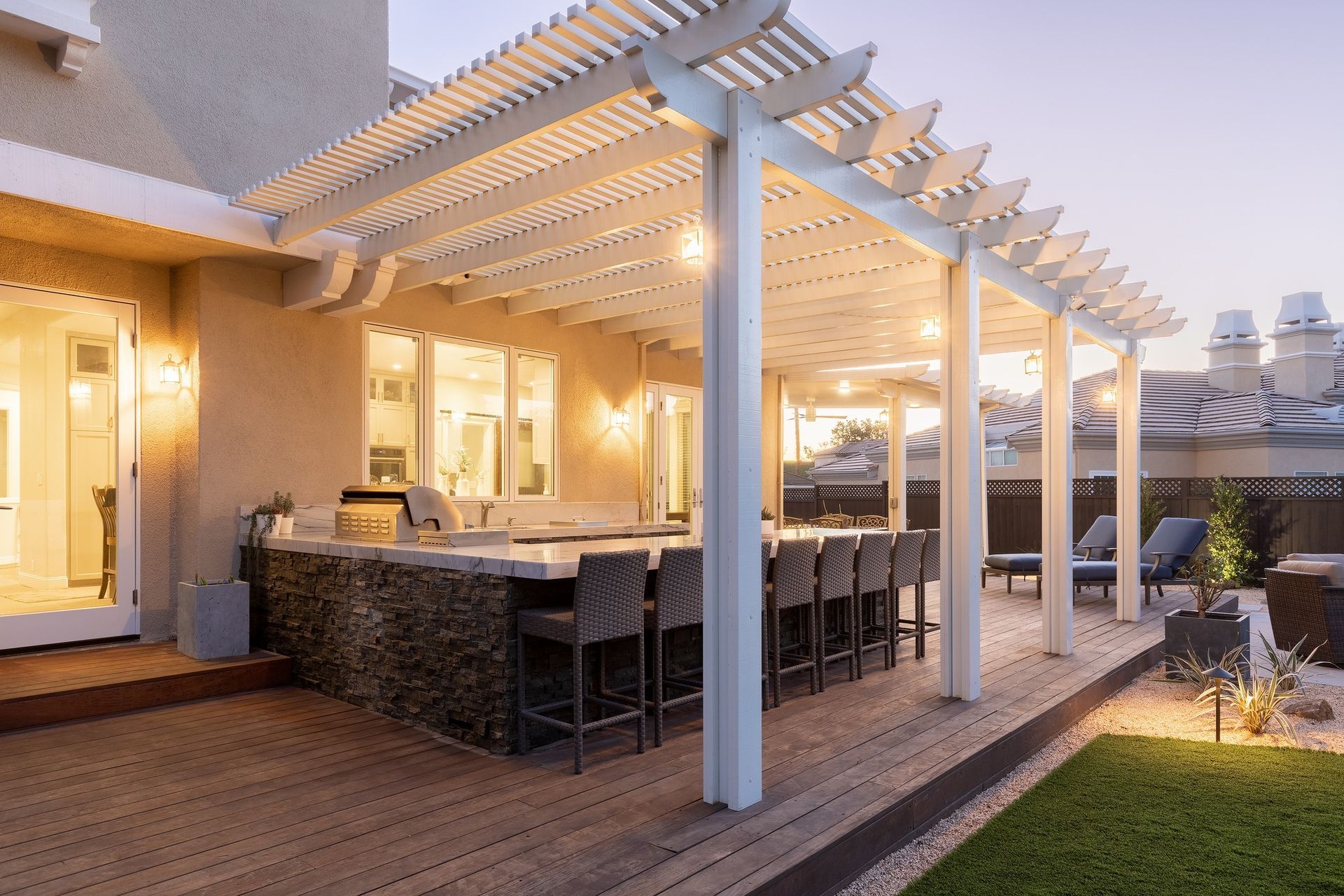 Backyard patio with white pergola, outdoor dining table, warm lights, and a grassy garden at dusk