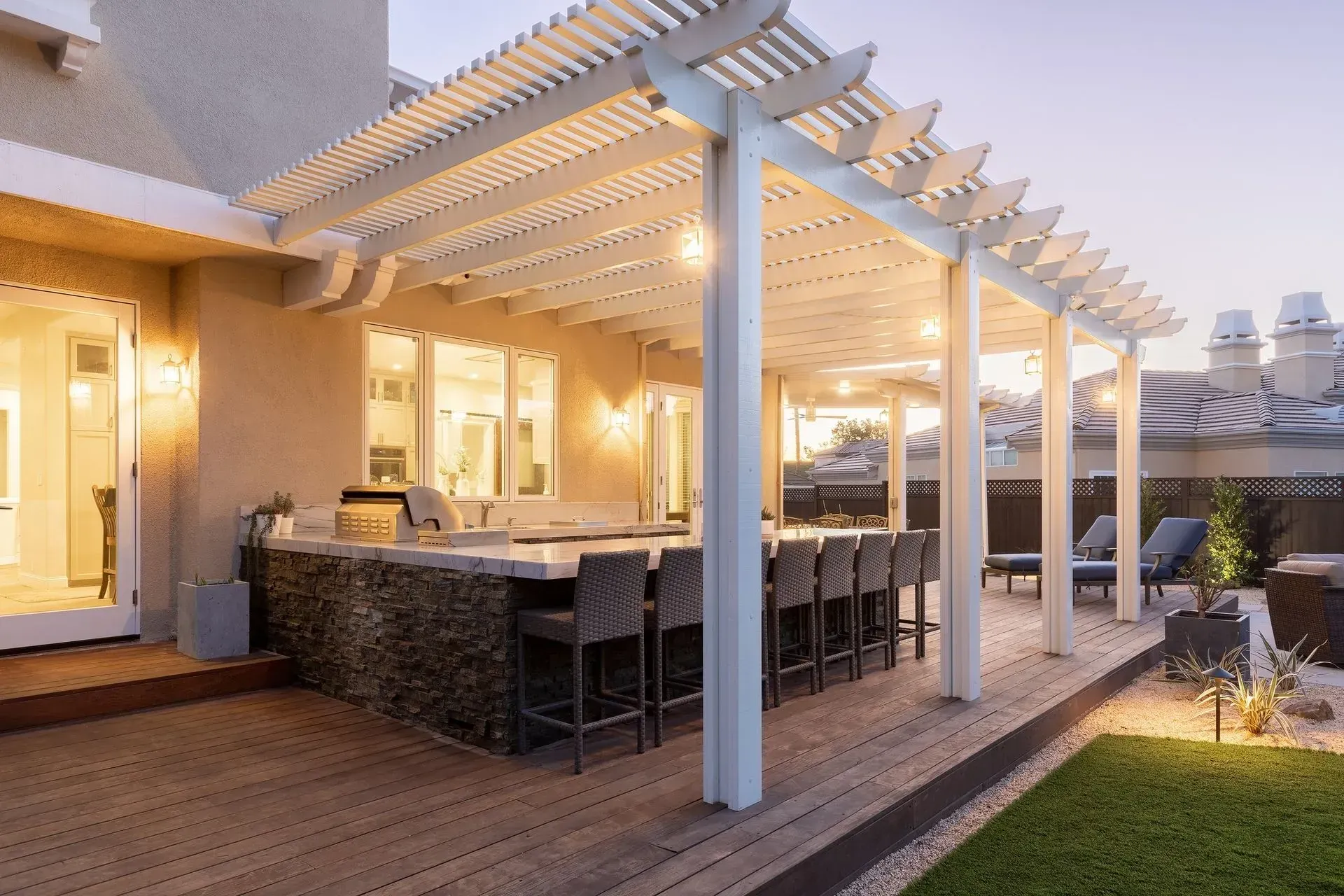 Modern outdoor patio with white pergola, bar seating, warm lights, and wooden deck at dusk