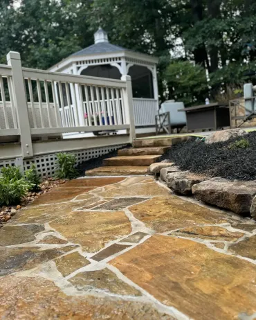 Stone walkway leading to a white gazebo and deck in a wooded backyard.