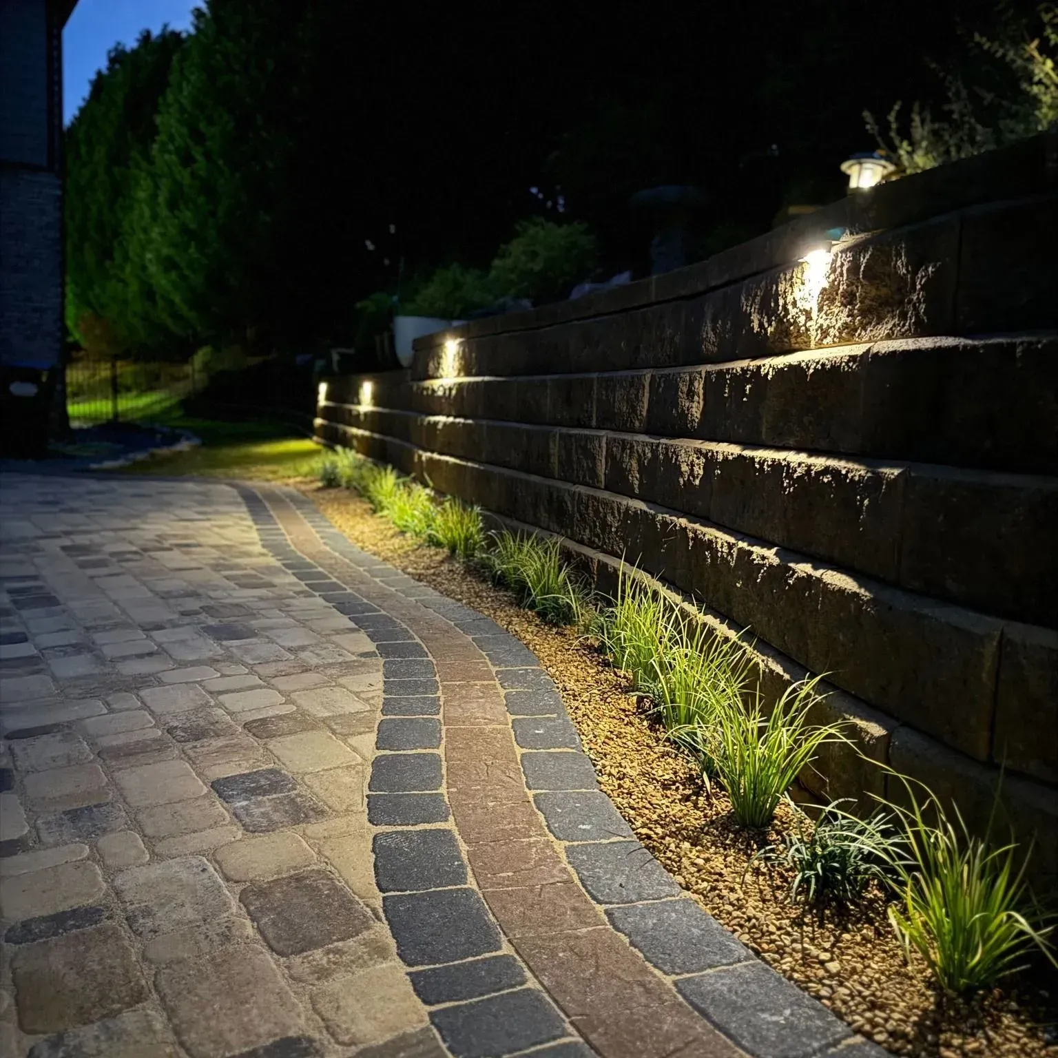 Curved stone walkway at night beside a lit retaining wall and grassy edge