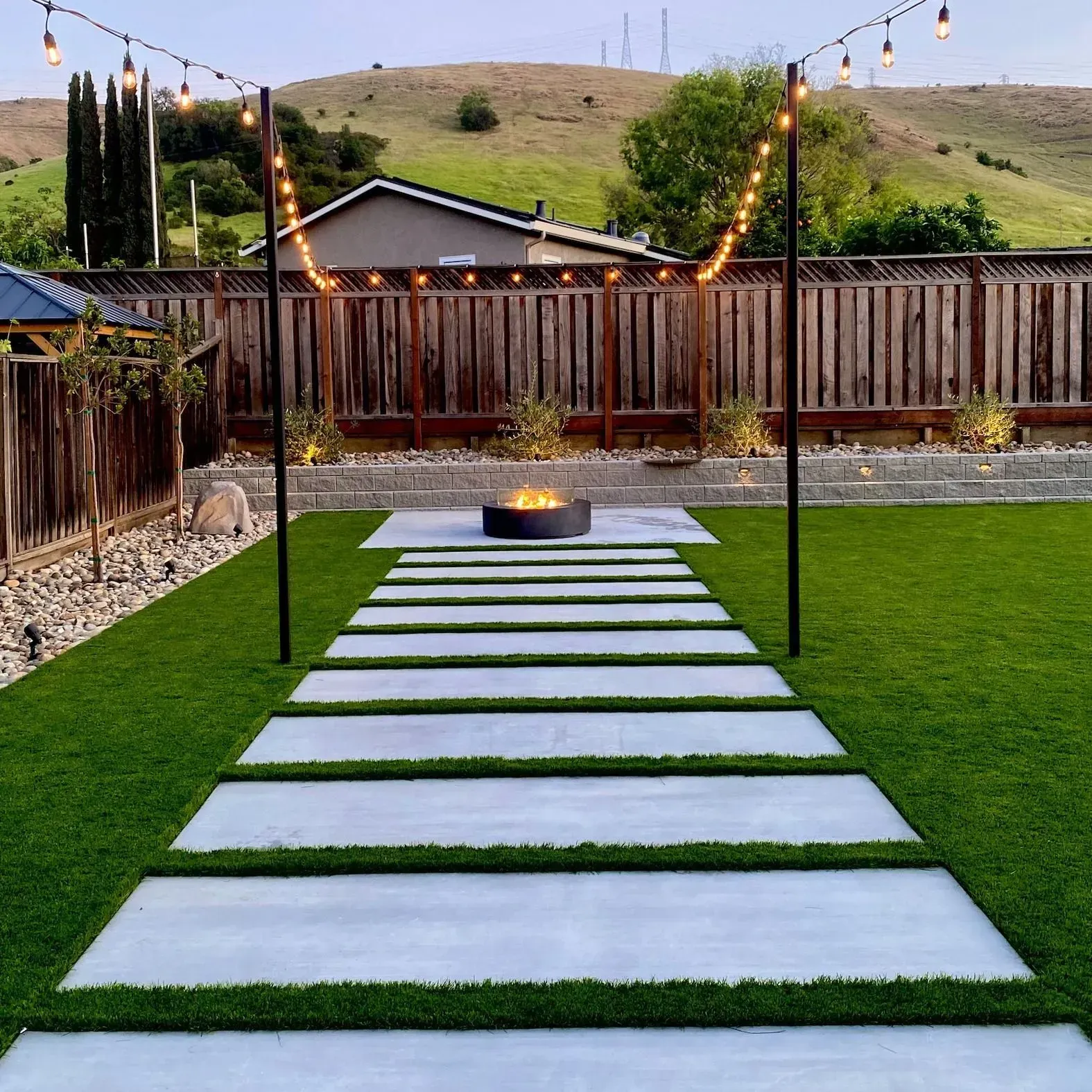 Backyard path of white stepping stones over grass, lit by string lights with a fire pit and fenced yard