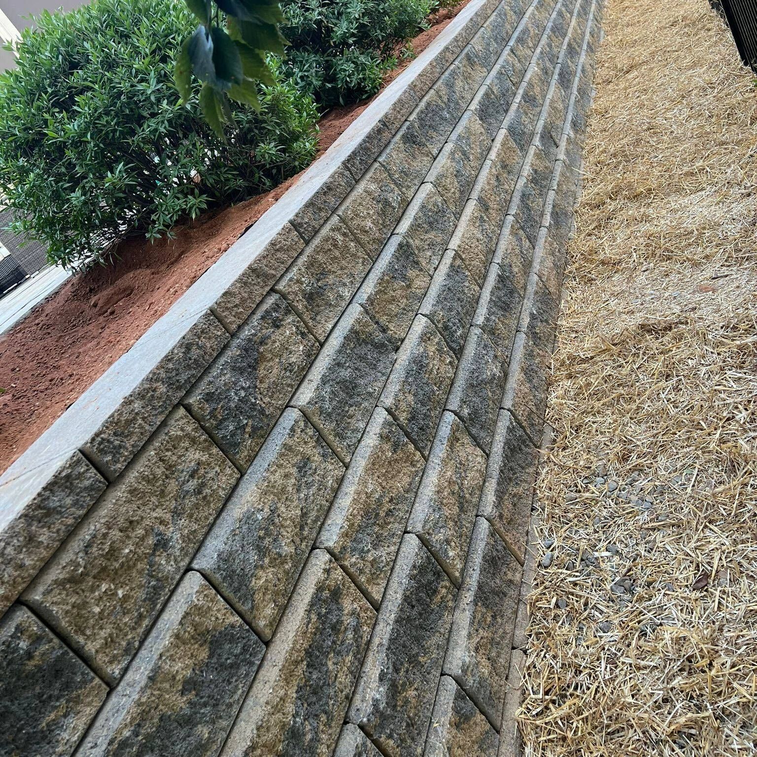 Angled view of a stone retaining wall beside shrubs and dry grass in a landscaped area