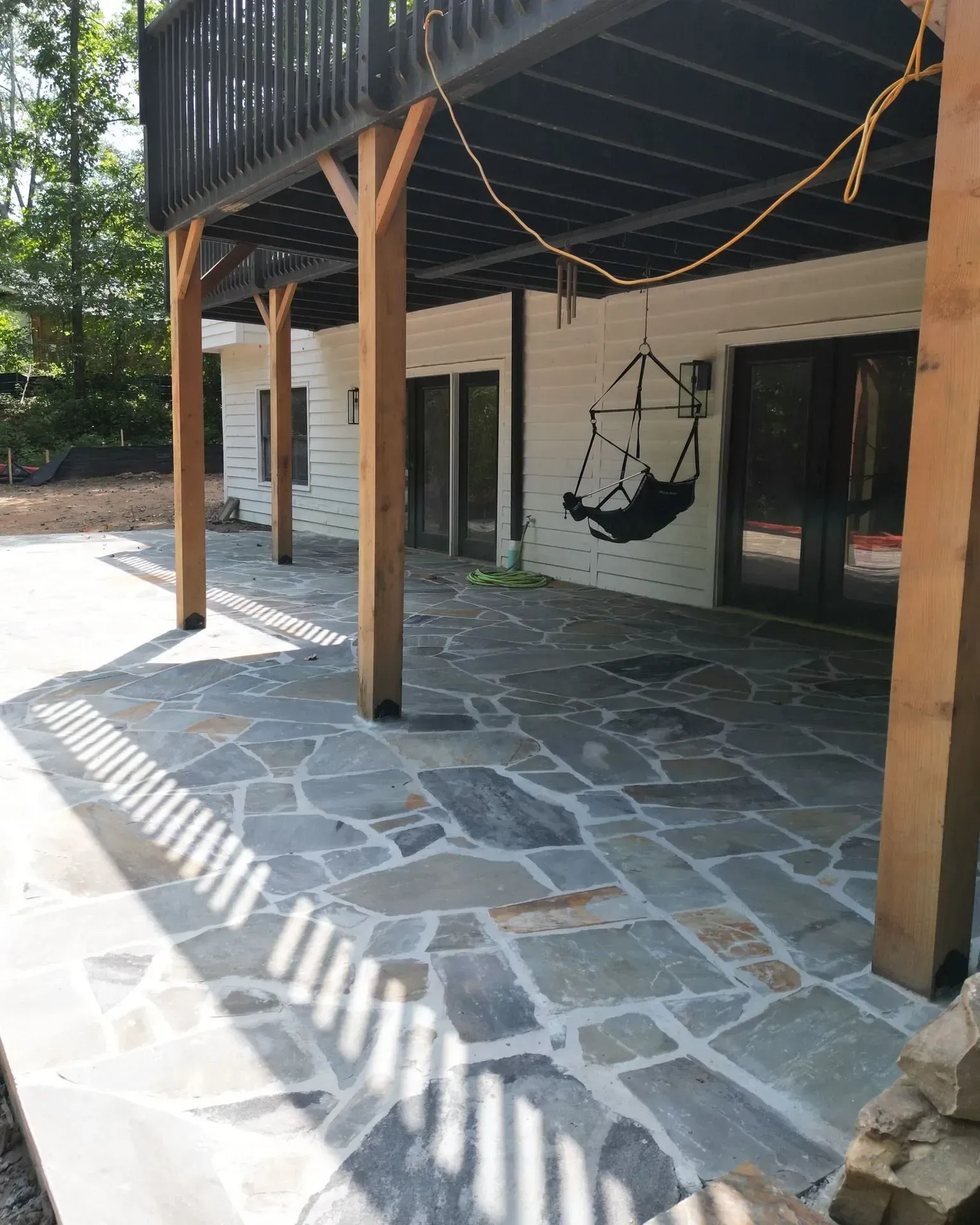 Stone patio under a wooden deck with a hanging chair beside a house exterior