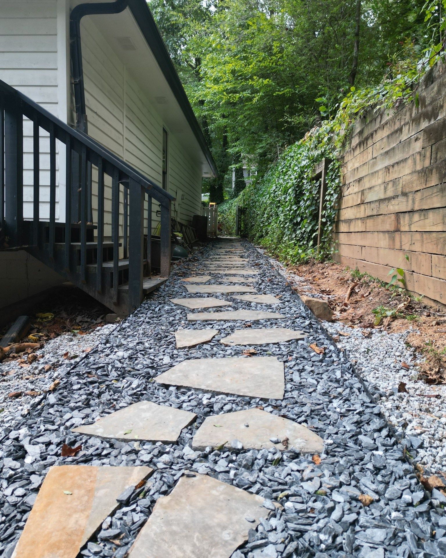 Stepping-stone path along a house beside a brick retaining wall, bordered by gravel and greenery