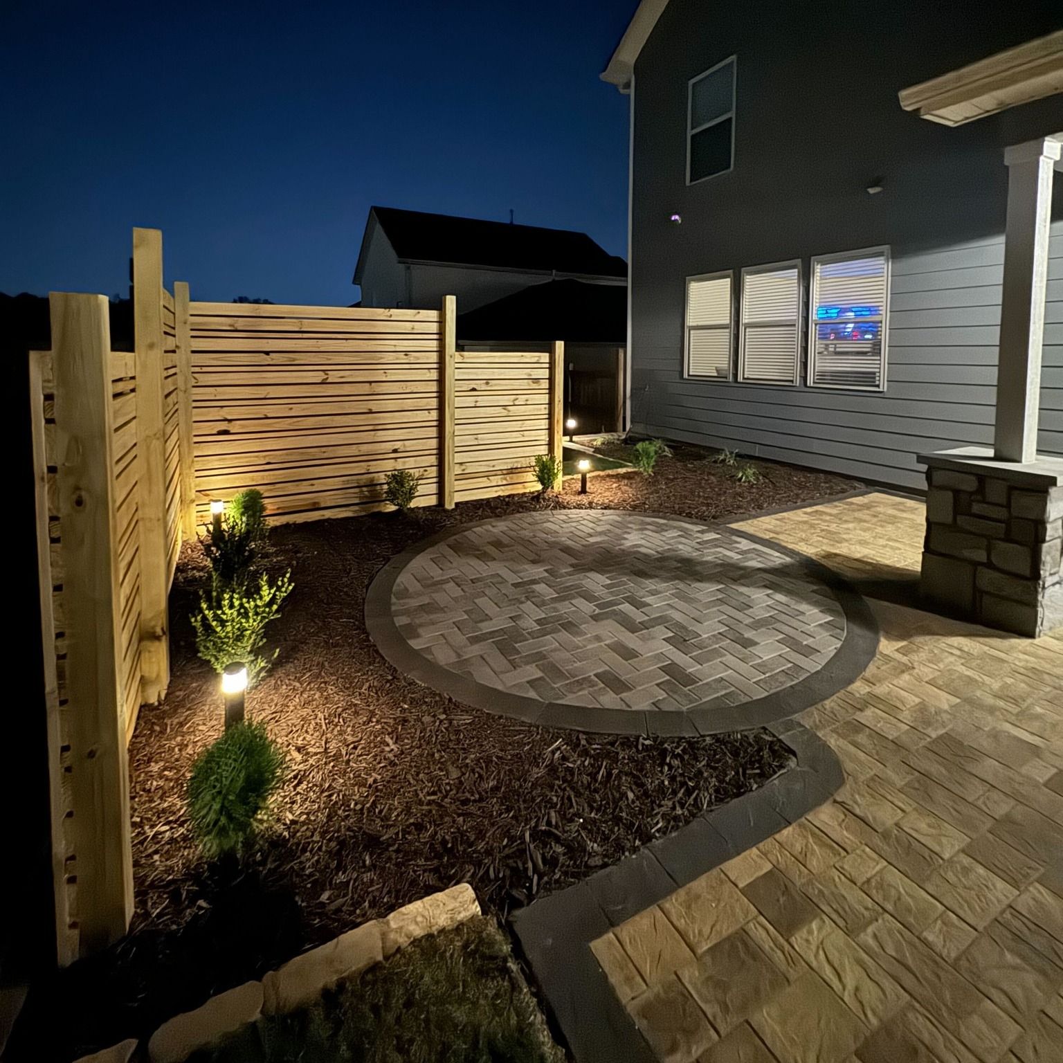 Backyard patio with circular stone fire pit, wooden fence, and lit landscaping beside a house at night