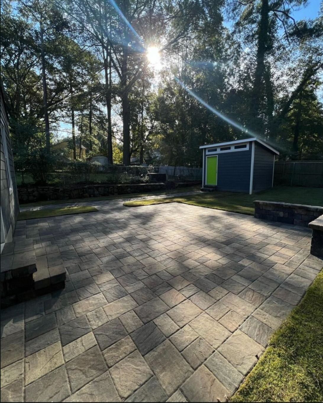 Sunny backyard patio with stone pavers, trees, and a small blue shed with a green door.