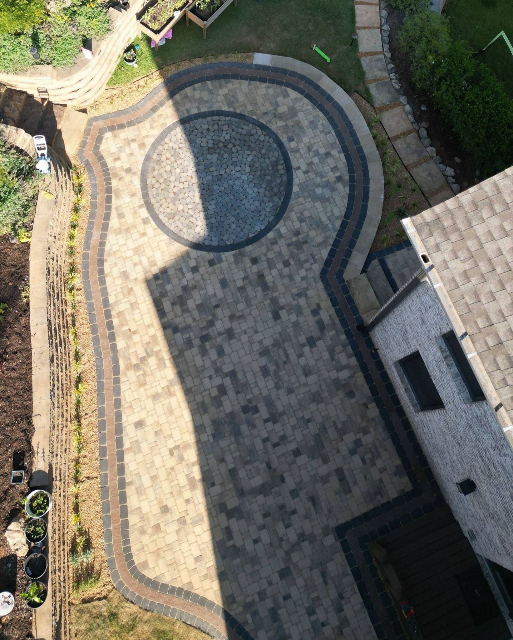 Aerial view of a curved stone patio beside a house, with a circular dark inlay and garden edging.