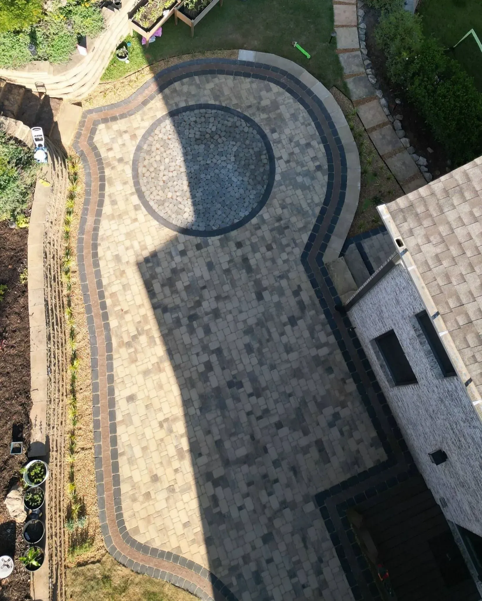 Aerial view of a paved stone patio with a curved dark border beside a house and garden.