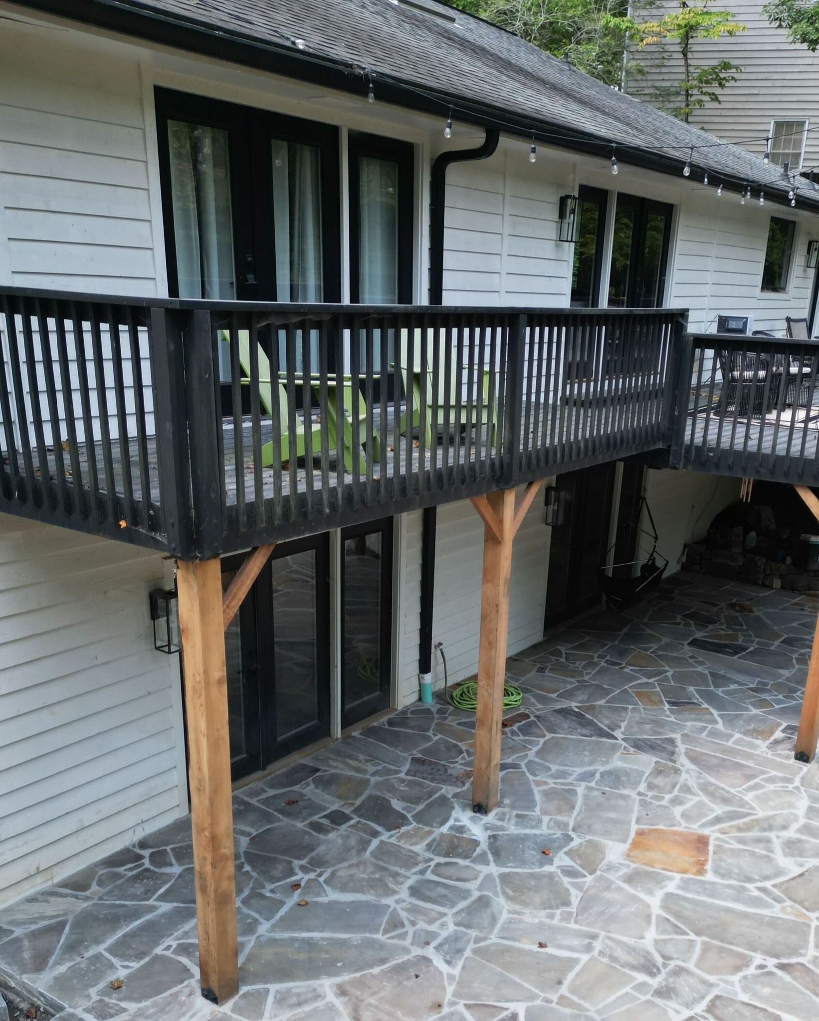 Wooden balcony with black railing above a stone patio beside a white house.