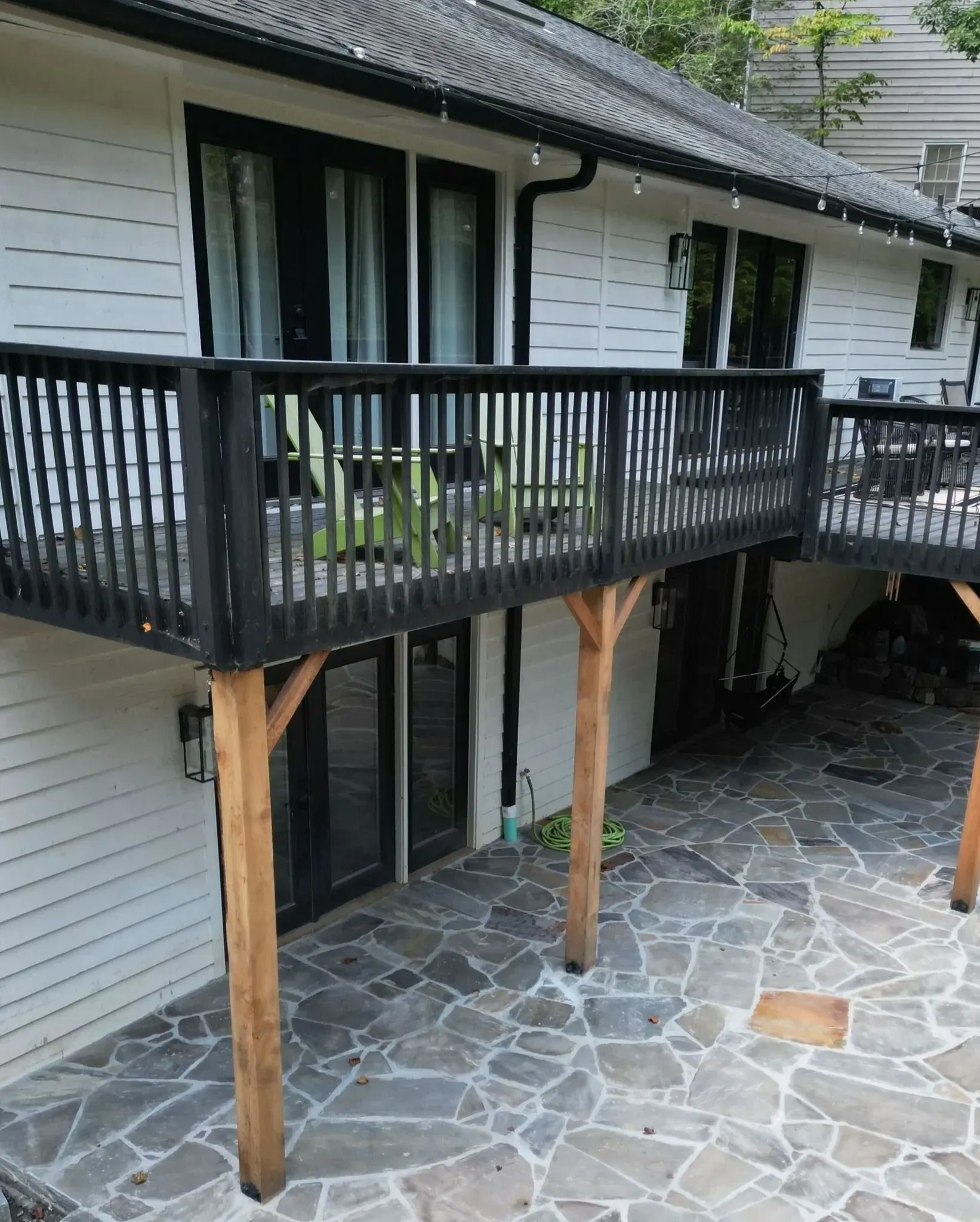Elevated deck with black railing beside a white house, supported by wooden posts over a stone patio.