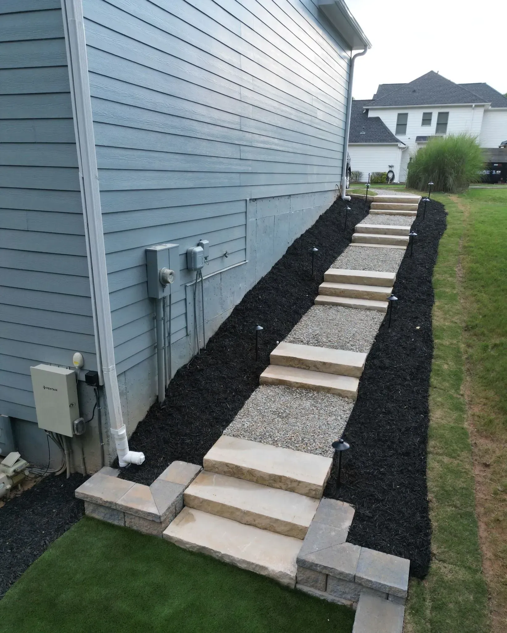 Stone walkway beside a blue house, bordered by dark mulch and leading across a green lawn.